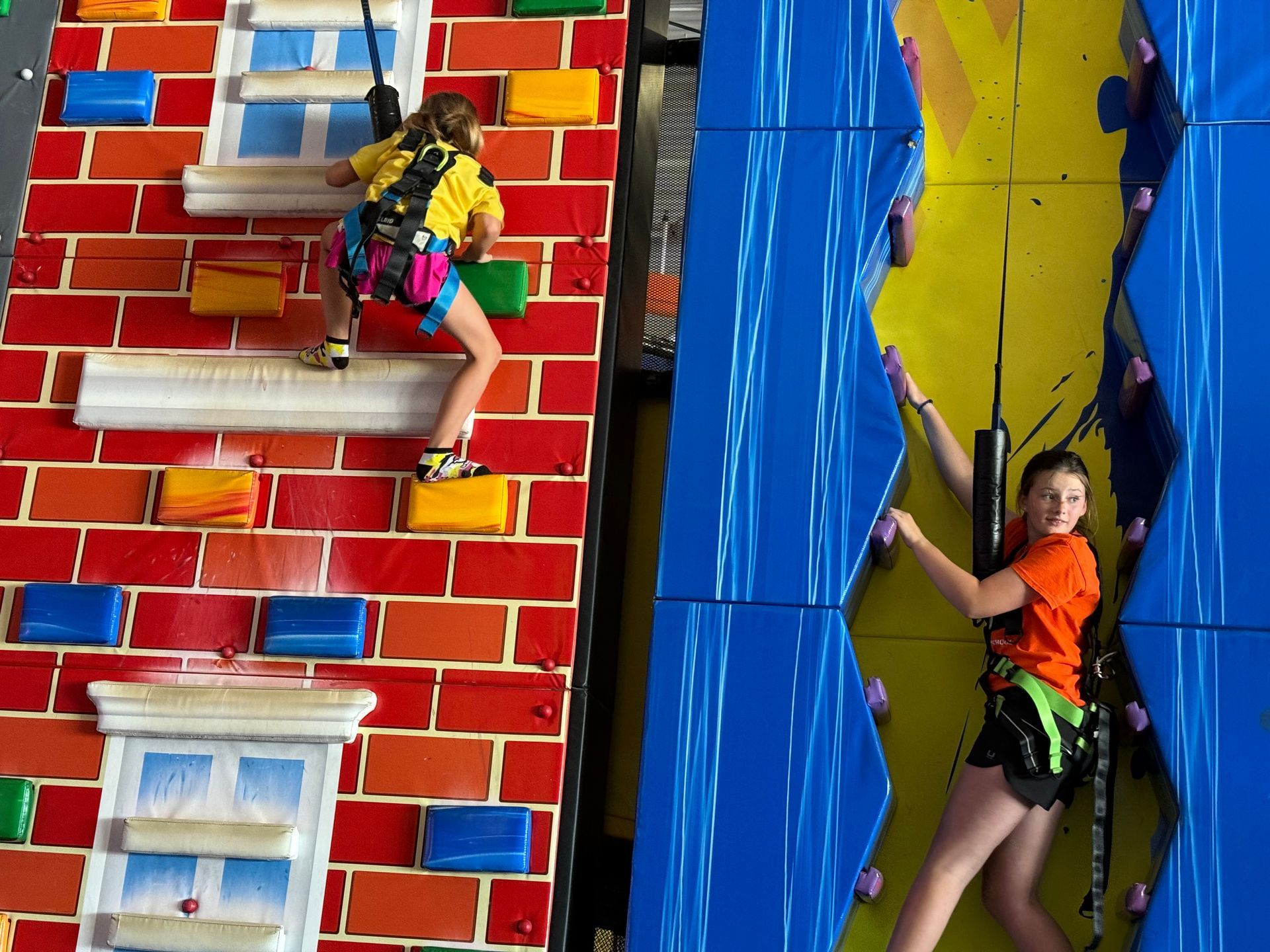 Two people climbing colorful indoor walls with harnesses and safety gear.