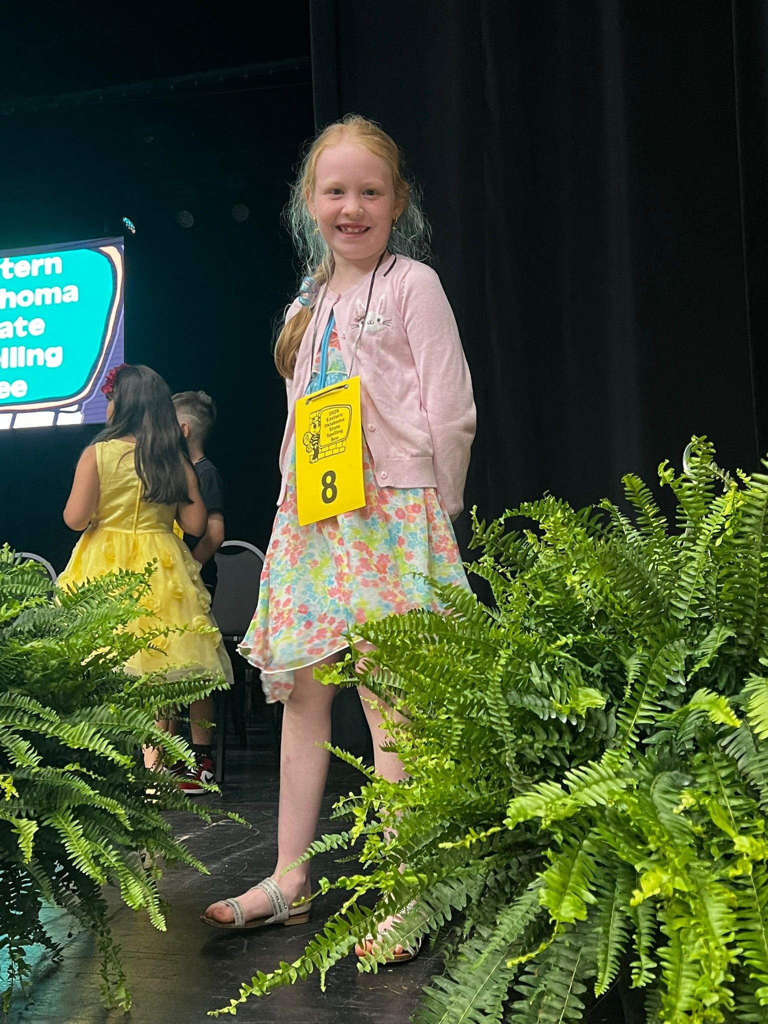 Girl on a stage smiling in a pink jacket and floral dress, with yellow flowers and ferns in front.