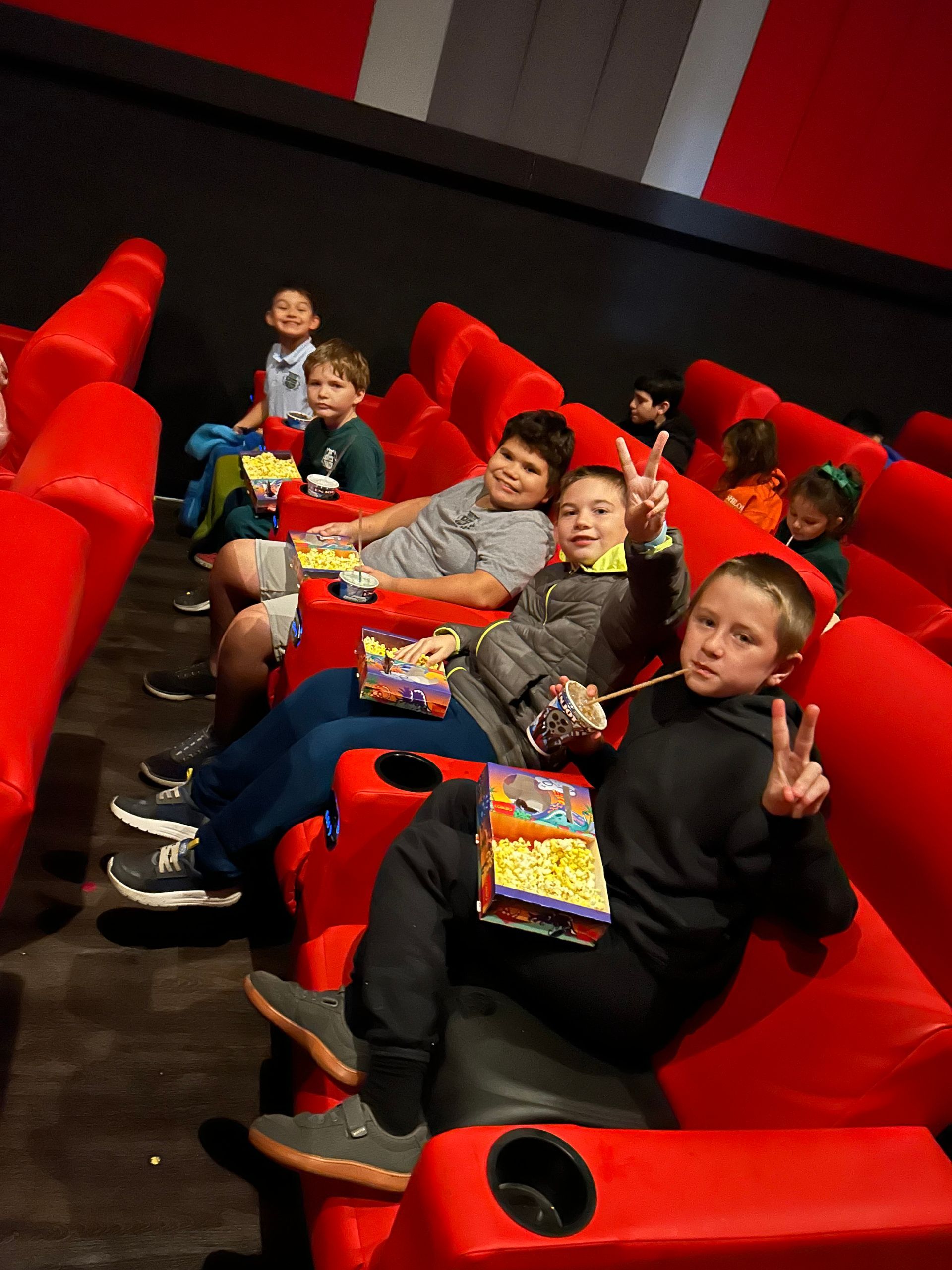 A group of children are sitting in red chairs in a movie theater eating popcorn.