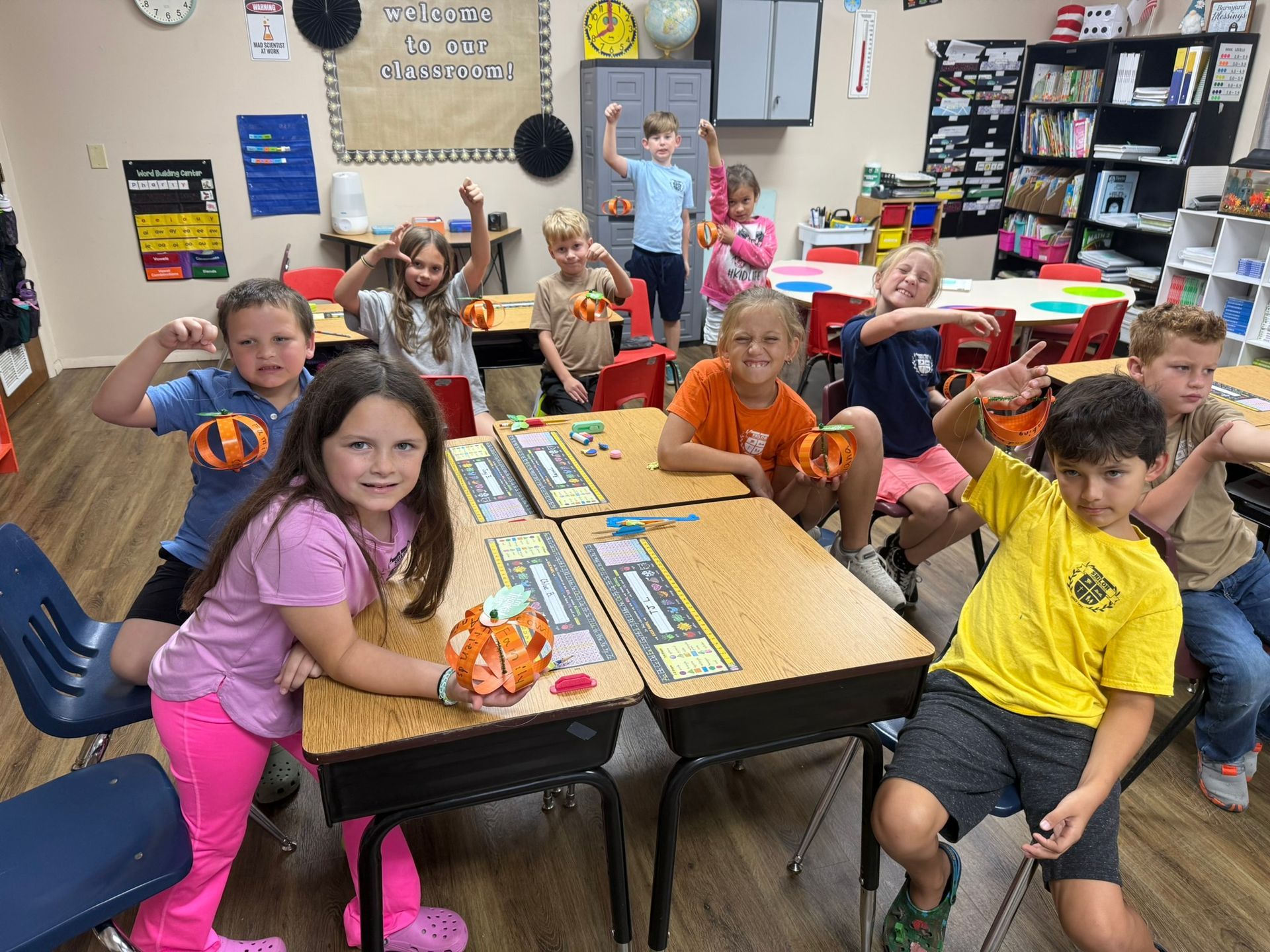 Children in a classroom pose with paper pumpkins, flexing muscles and smiling. Desks, books, and decorations are visible.