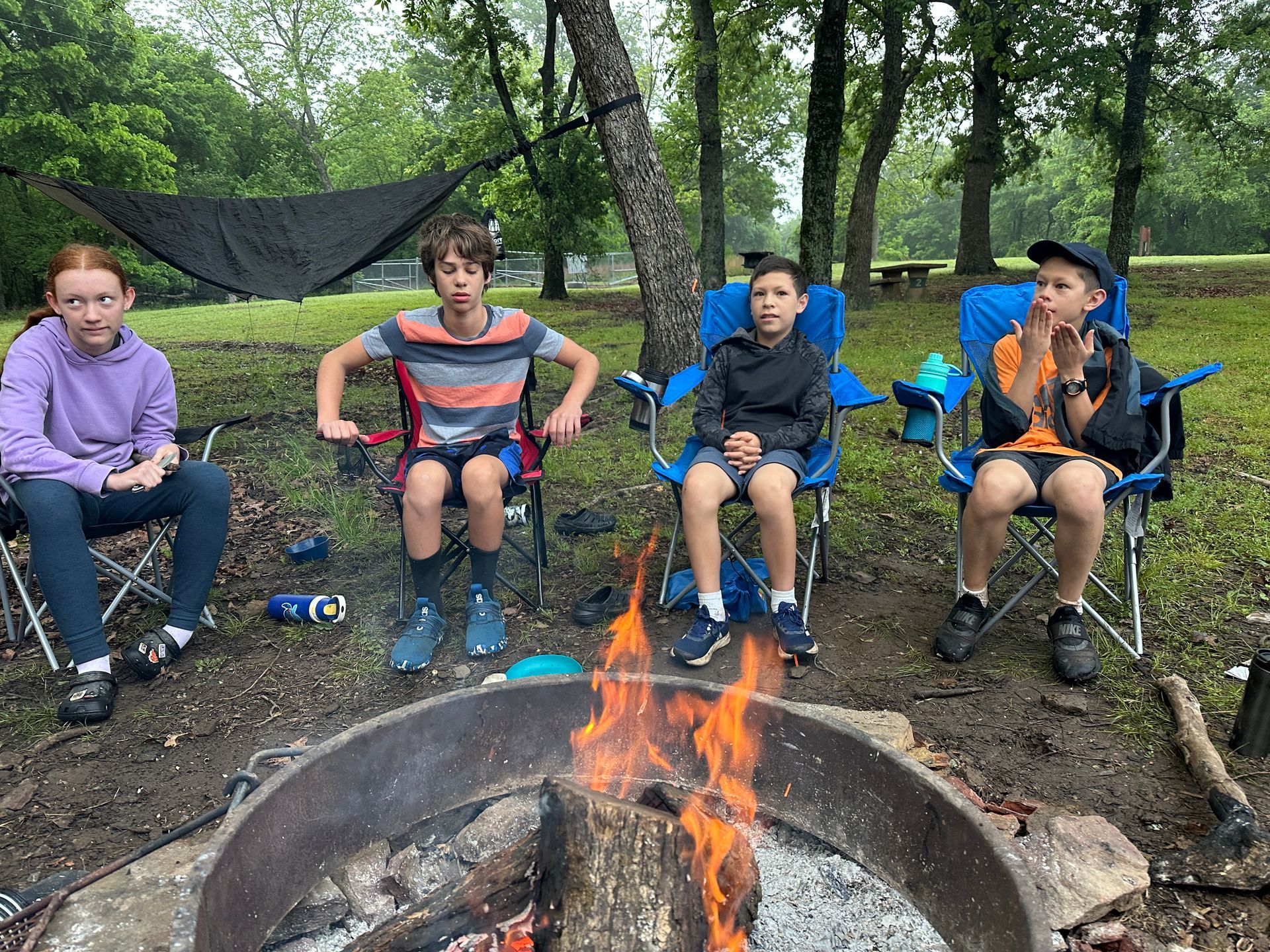 People seated around a campfire in a wooded campsite, with camping chairs and trees in the background.