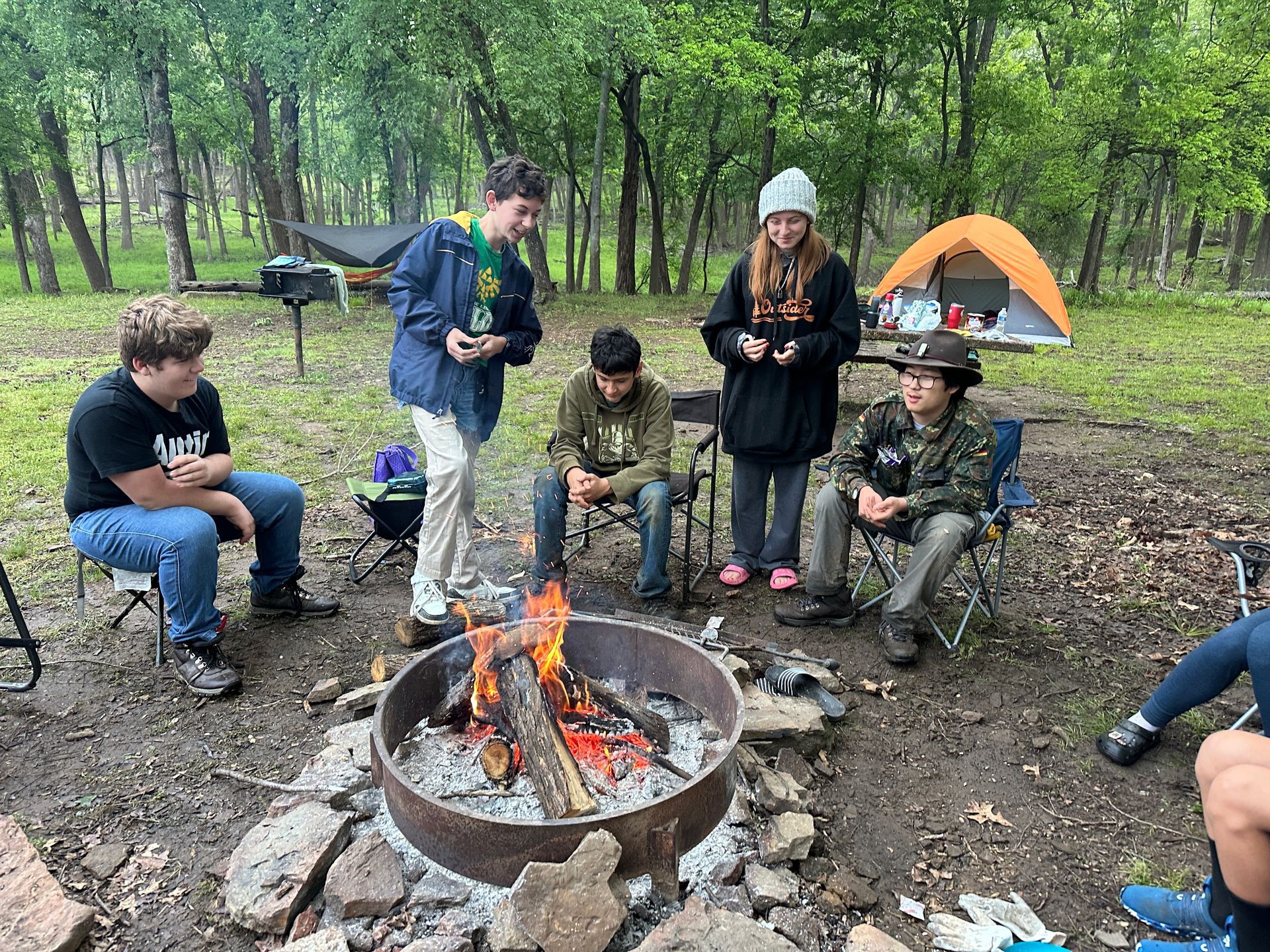 People gathered around a campfire in a wooded campsite, with tents and trees in the background.