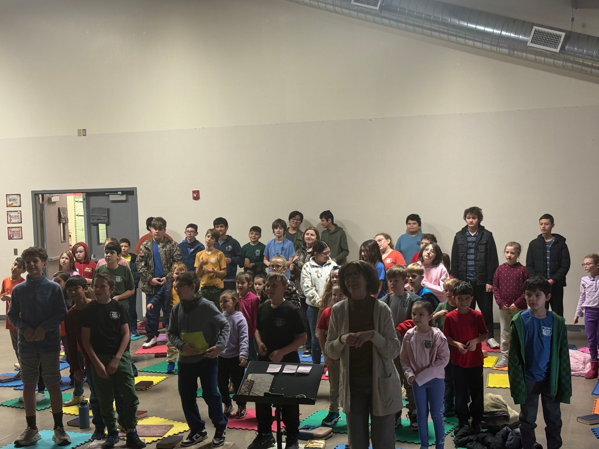 Large group of children and an adult in a large room, potentially for a performance or rehearsal.