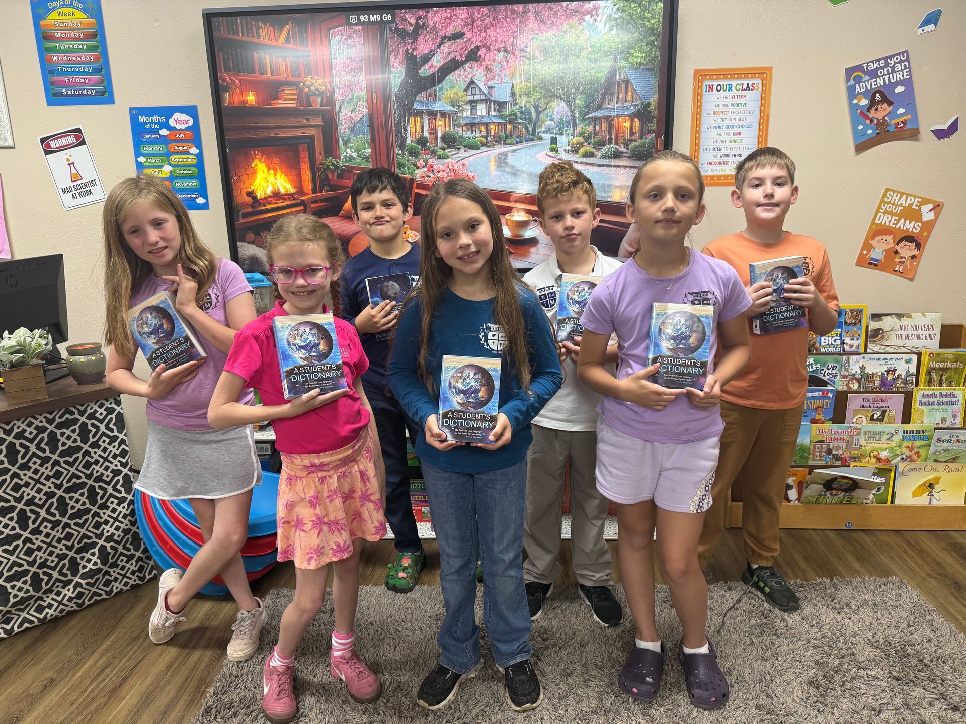Six children stand holding cups in a colorful classroom with a fireplace mural and posters.