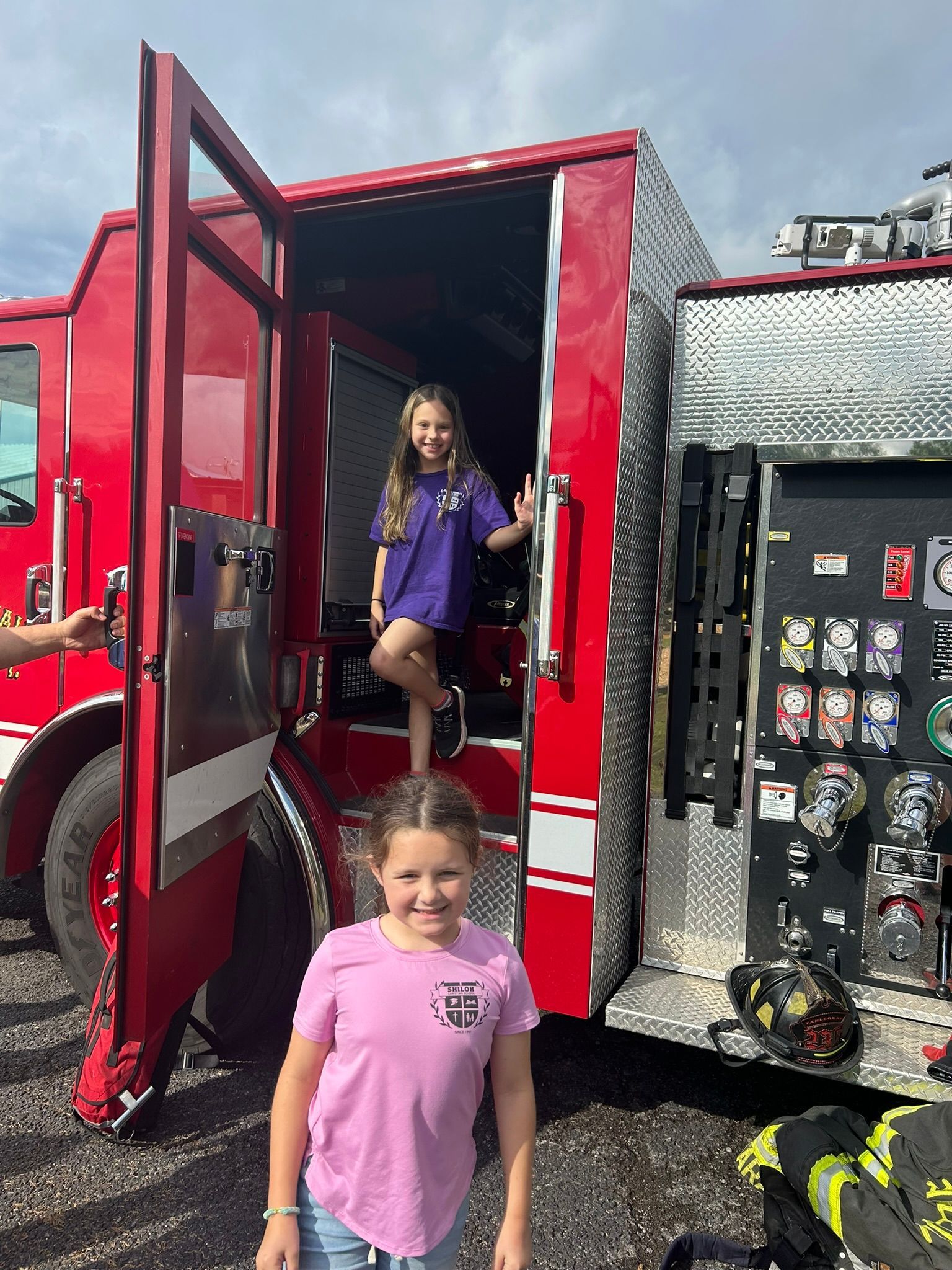 Two young girls pose near a red fire truck. One stands in the doorway, the other in front, both smiling.