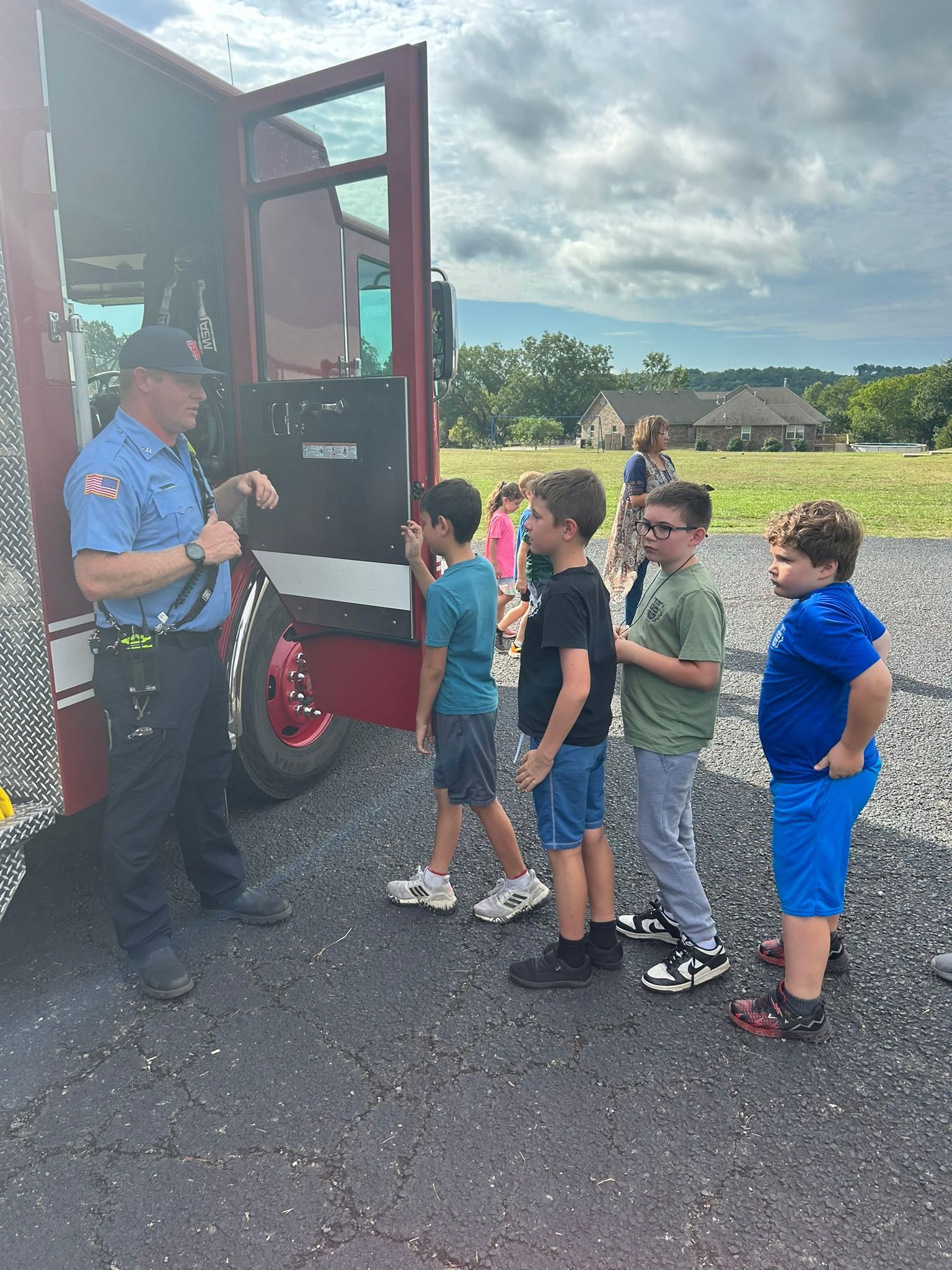 Firefighter speaking to a group of children near a fire truck on a cloudy day.