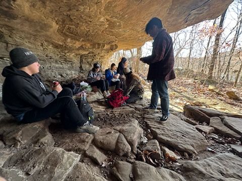 A group of people are sitting under a cave in the woods.