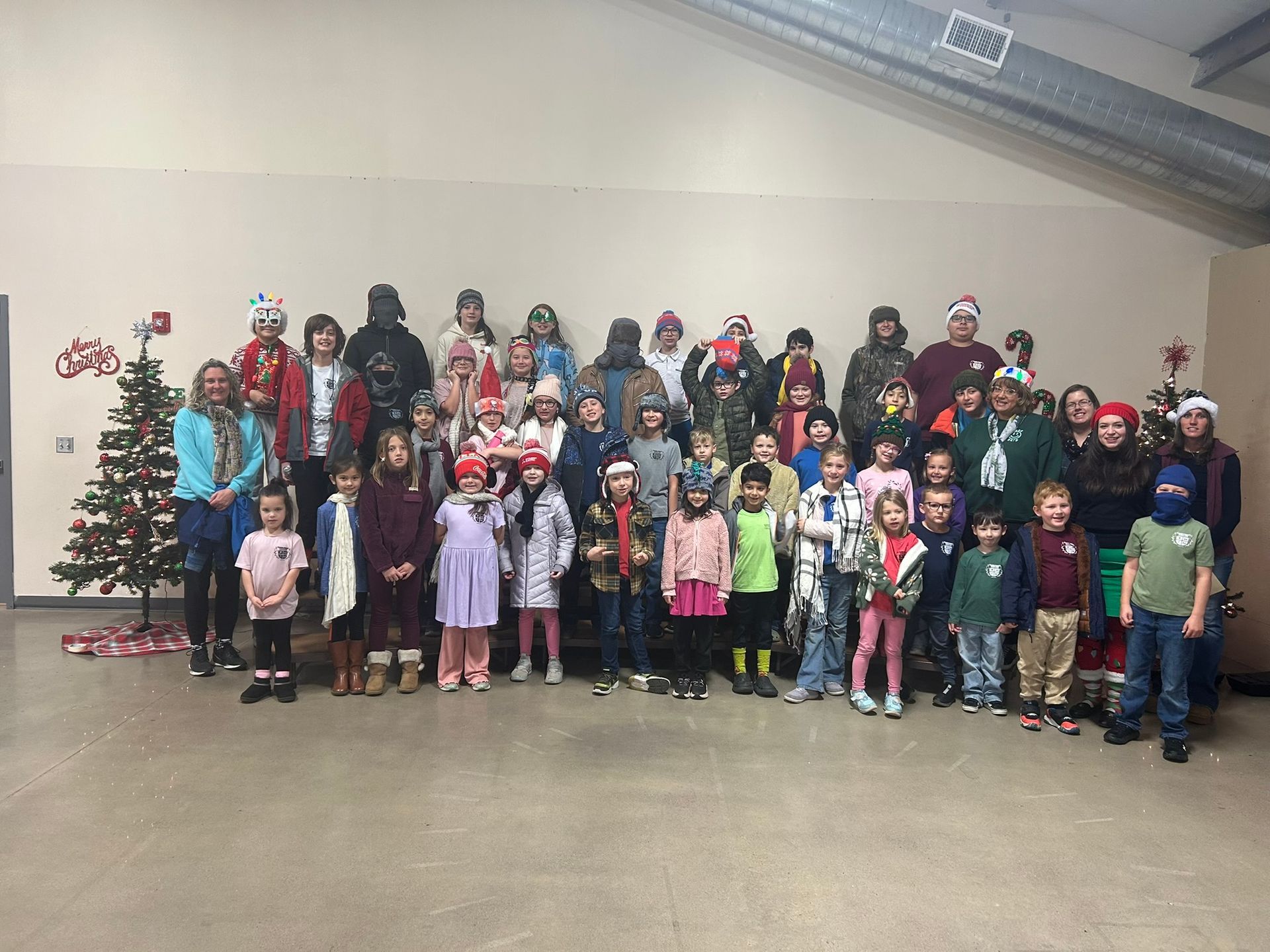 Group of people, mostly children, posing for photo in room with Christmas decorations. Many wearing holiday hats.