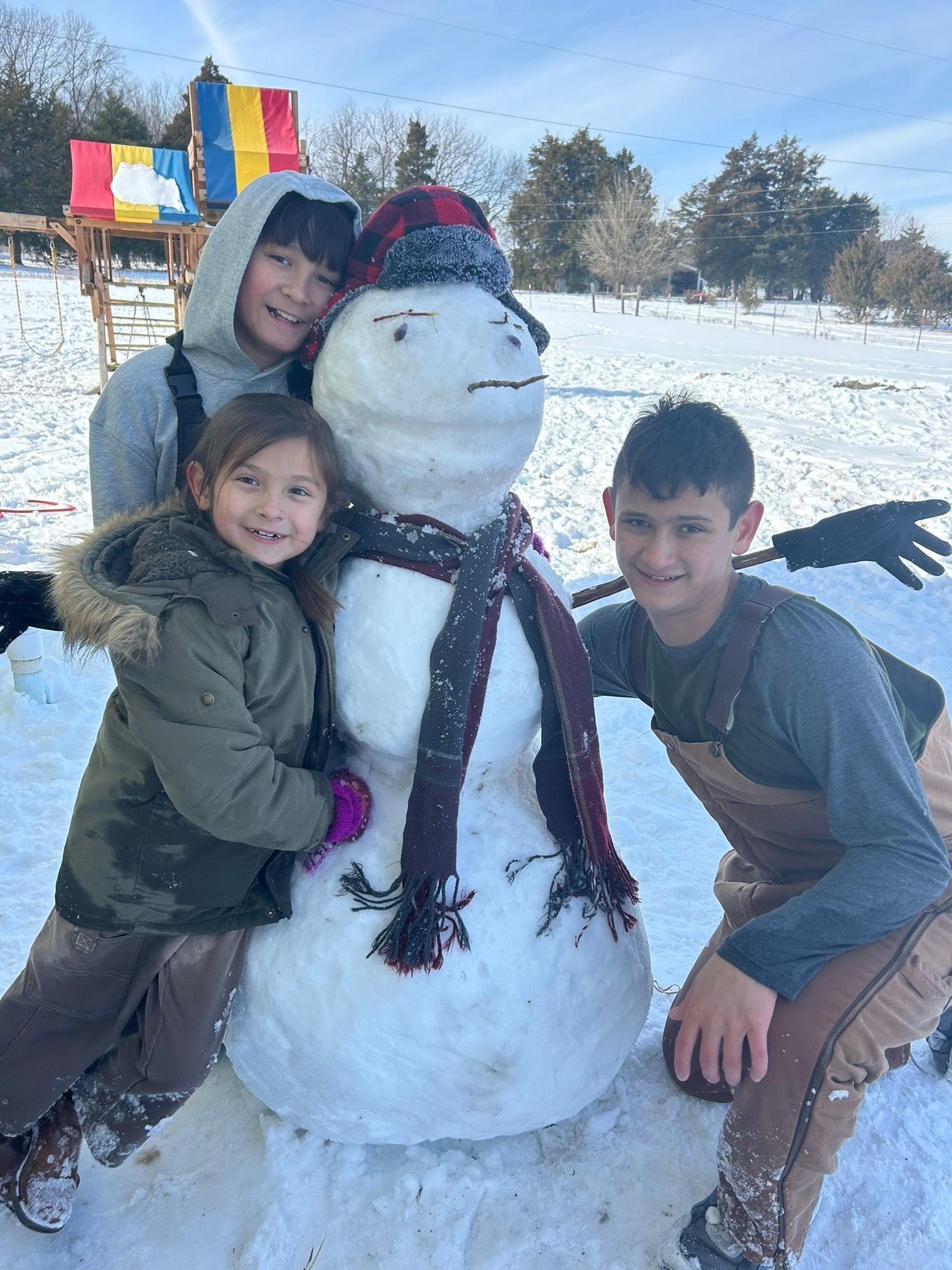 Three children pose with a snowman in a snowy outdoor setting.