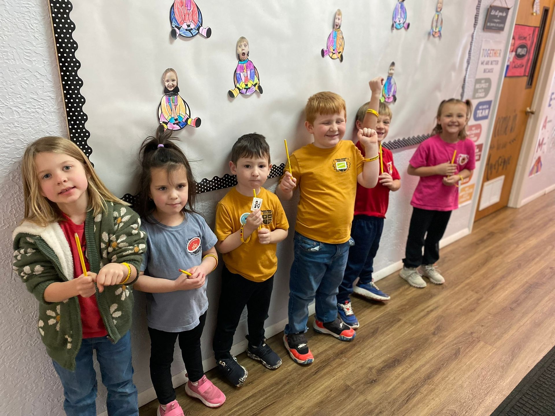 Children holding pencils stand in front of a classroom banner with colorful drawings.