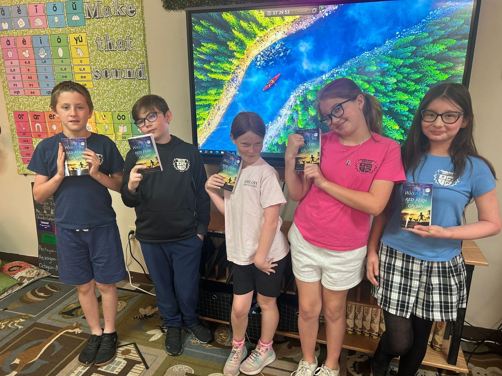 Five children stand in a classroom holding small items in front of a bright aquarium mural.