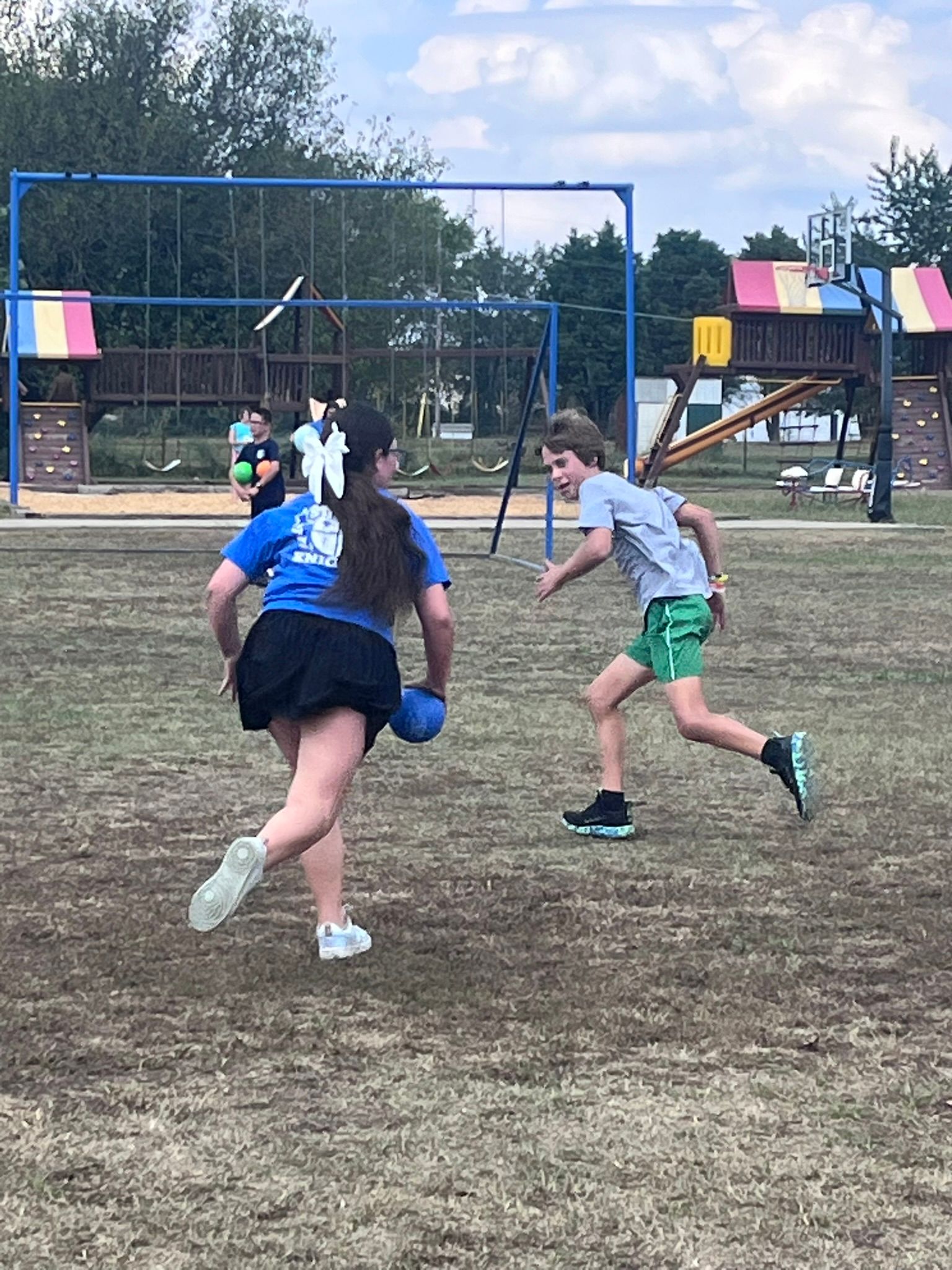 Two children playing dodgeball on a grassy field. The girl is wearing a blue shirt, and the boy is wearing a gray shirt.