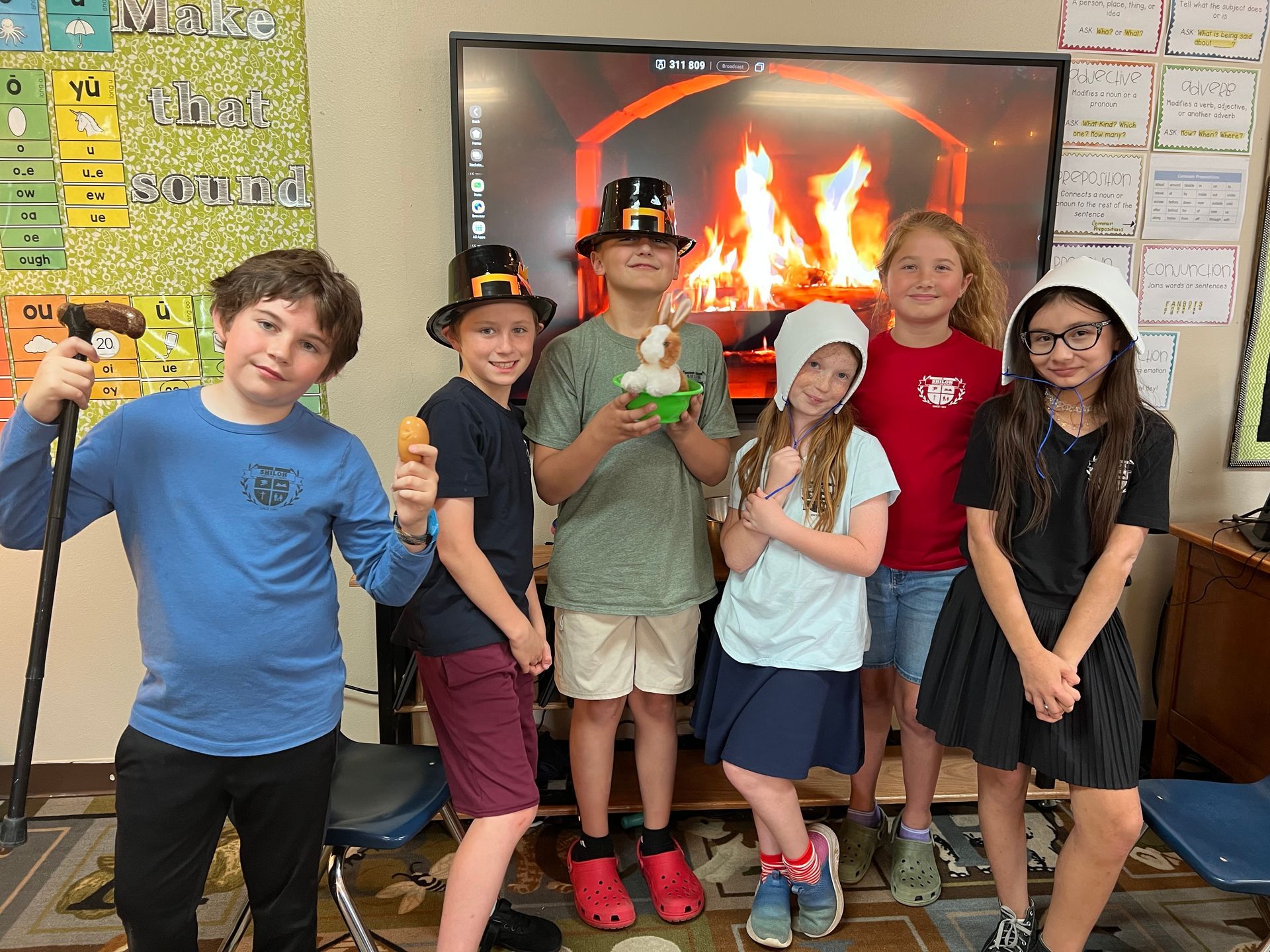 Group of children in hats posing in a classroom with a fireplace backdrop.
