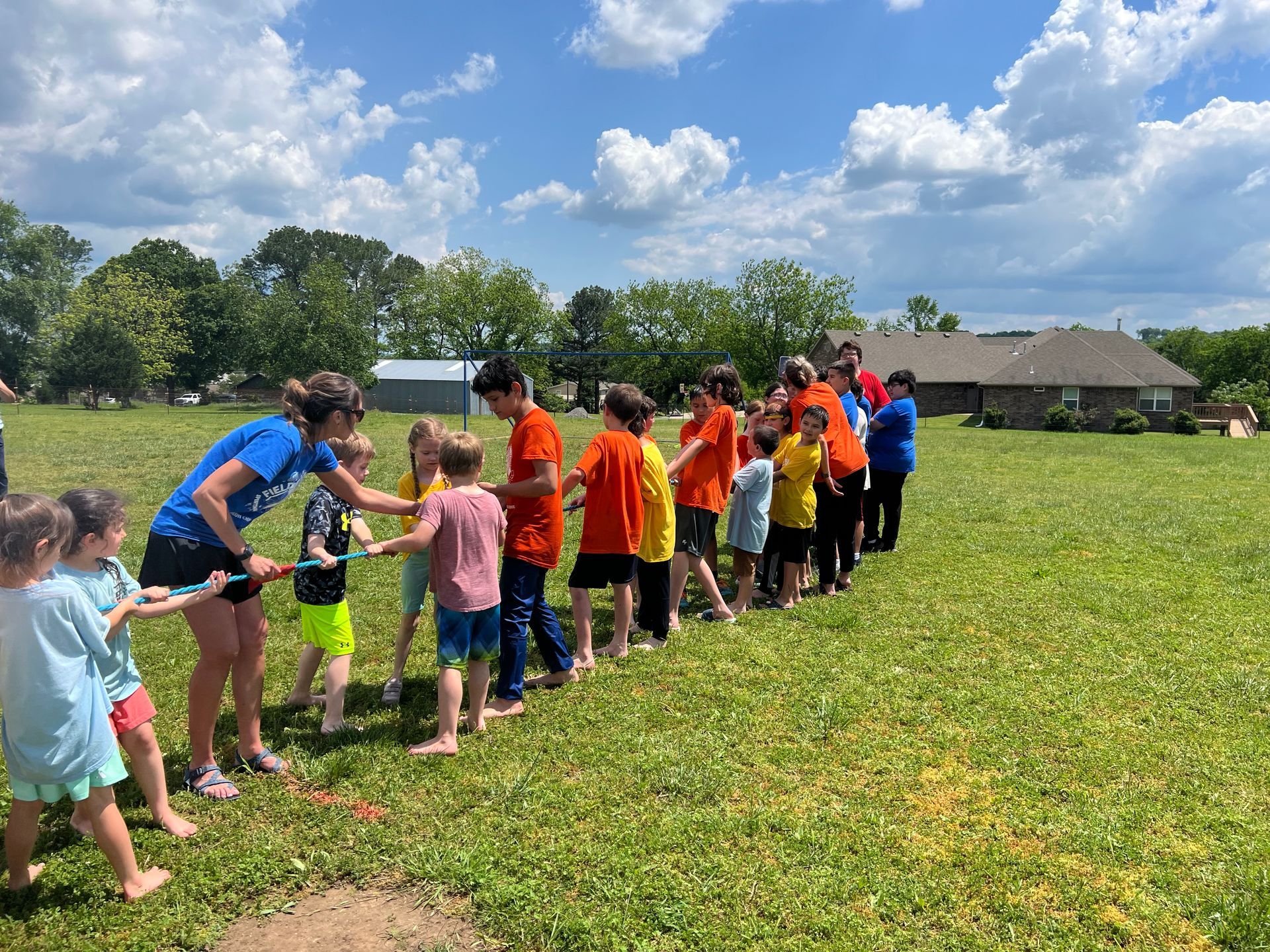 Children playing tug-of-war on a grassy field under a blue sky. One team is blue/white and the other orange/yellow.