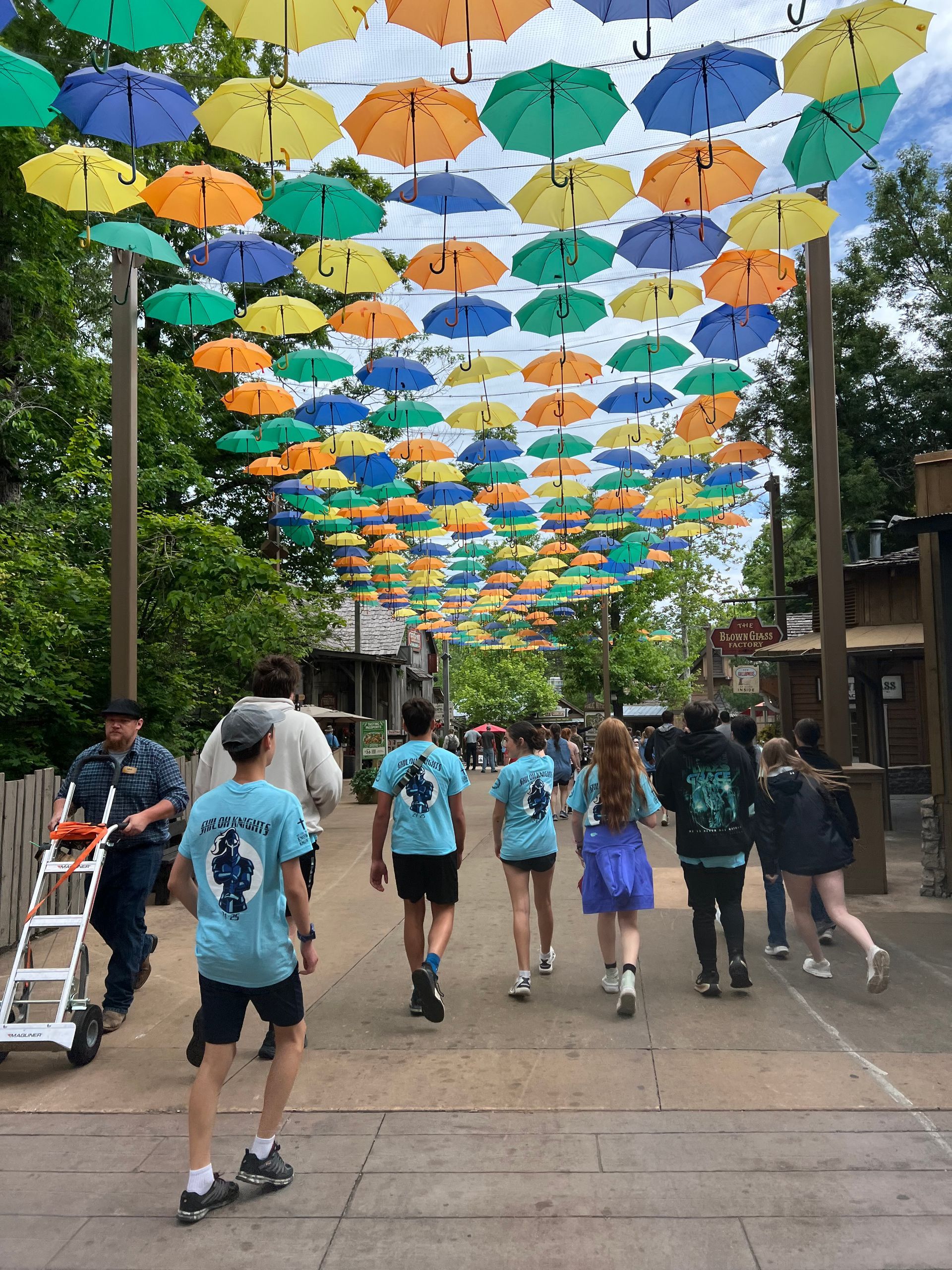 People walk beneath a canopy of colorful umbrellas on a street in a sunny outdoor setting.
