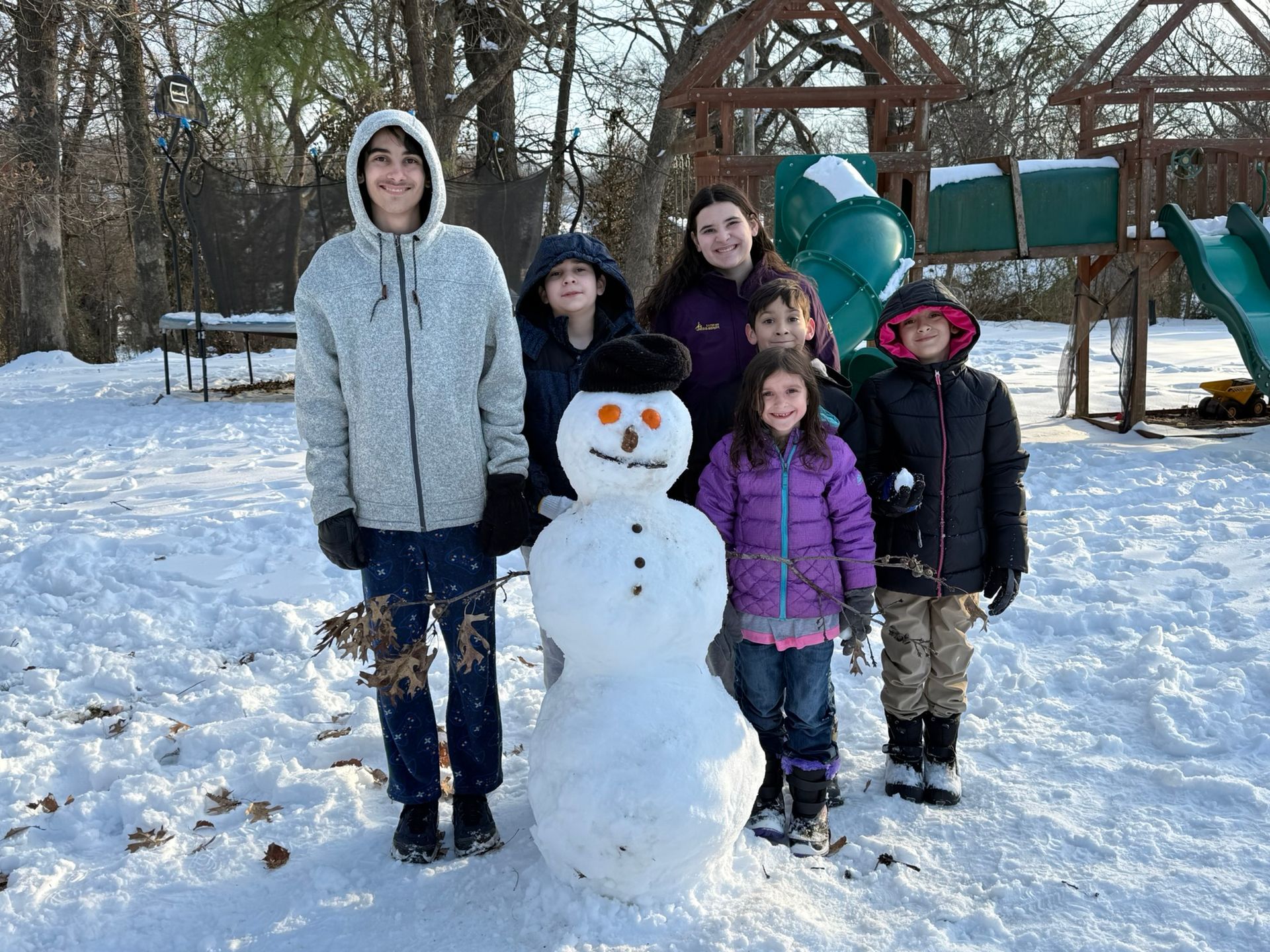 Group of children standing with a snowman in a snowy yard, smiles. Playground in the background.