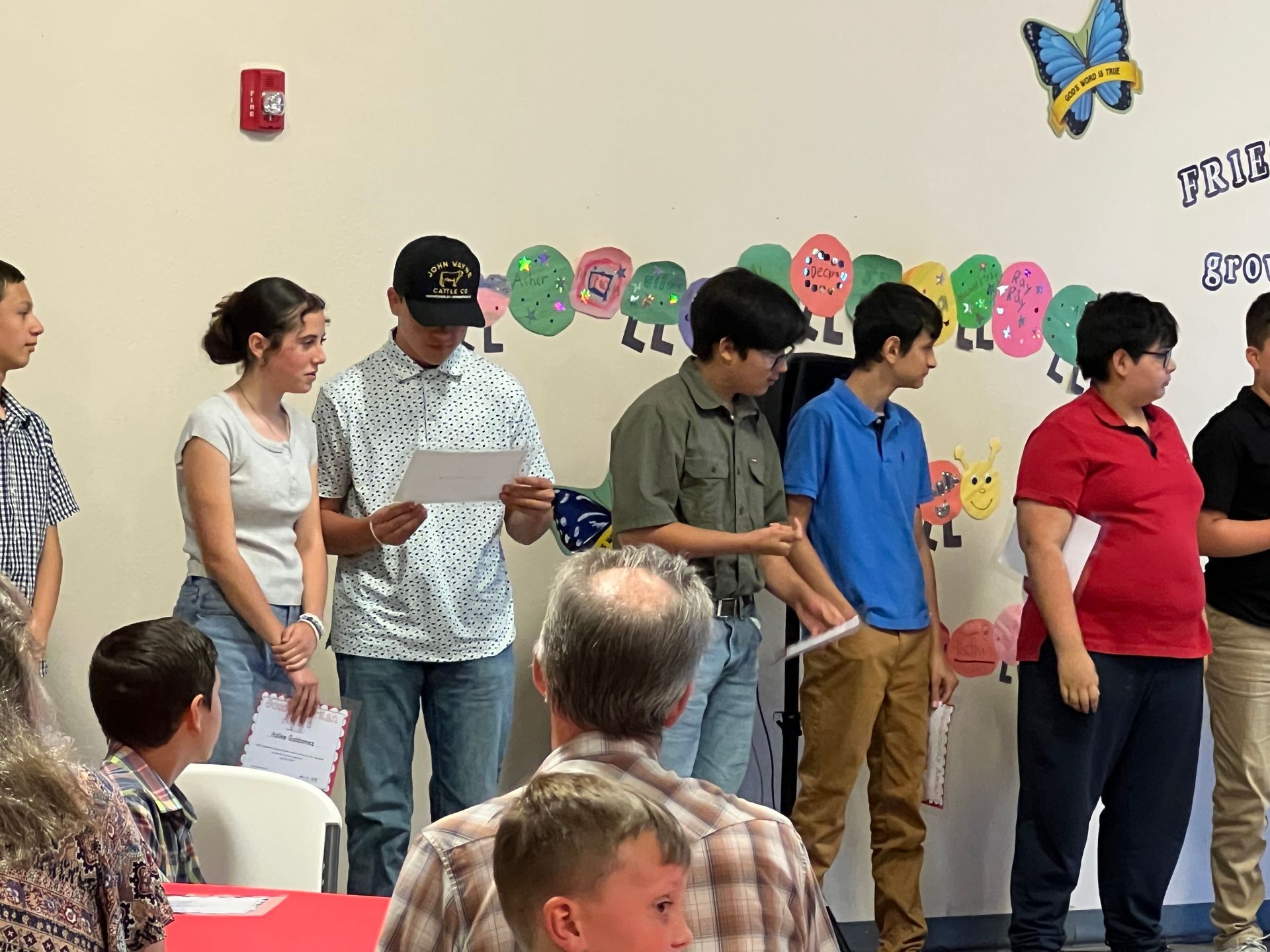 Group of students standing on a stage, some holding papers. A backdrop of caterpillar decor is visible.