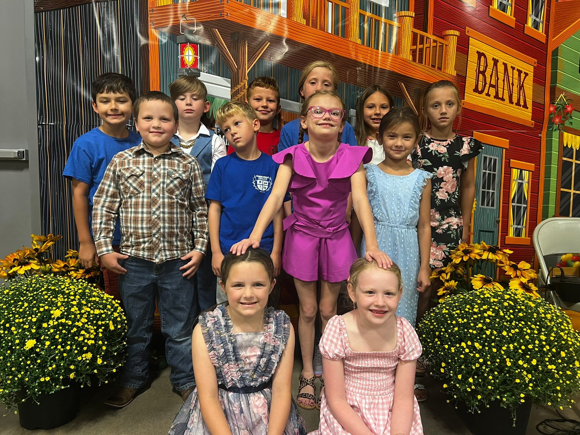 Group of children smiling at a backdrop of a Western town, decorated with fall flowers.