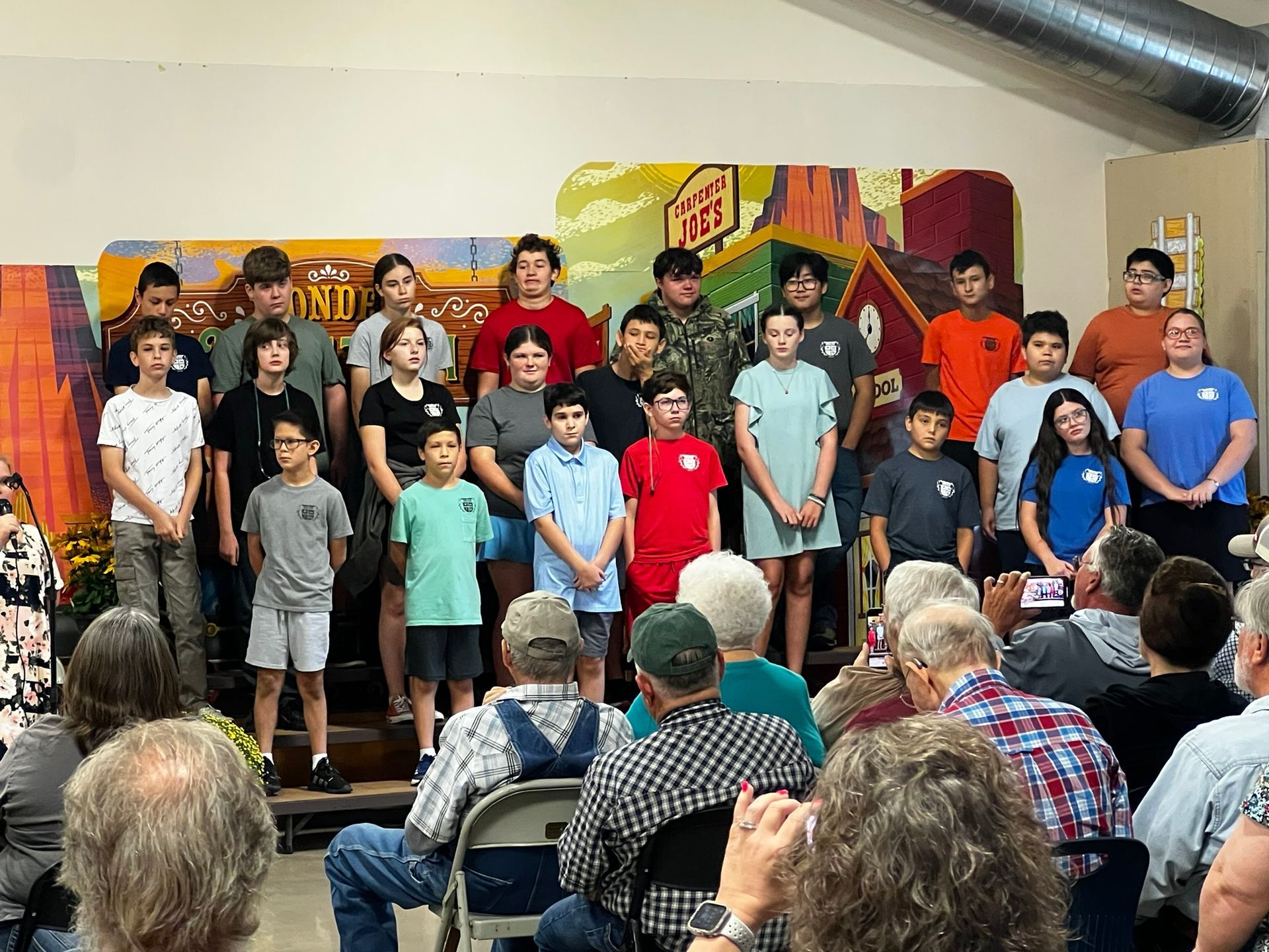 A group of teens and children stand on a stage in front of an audience.