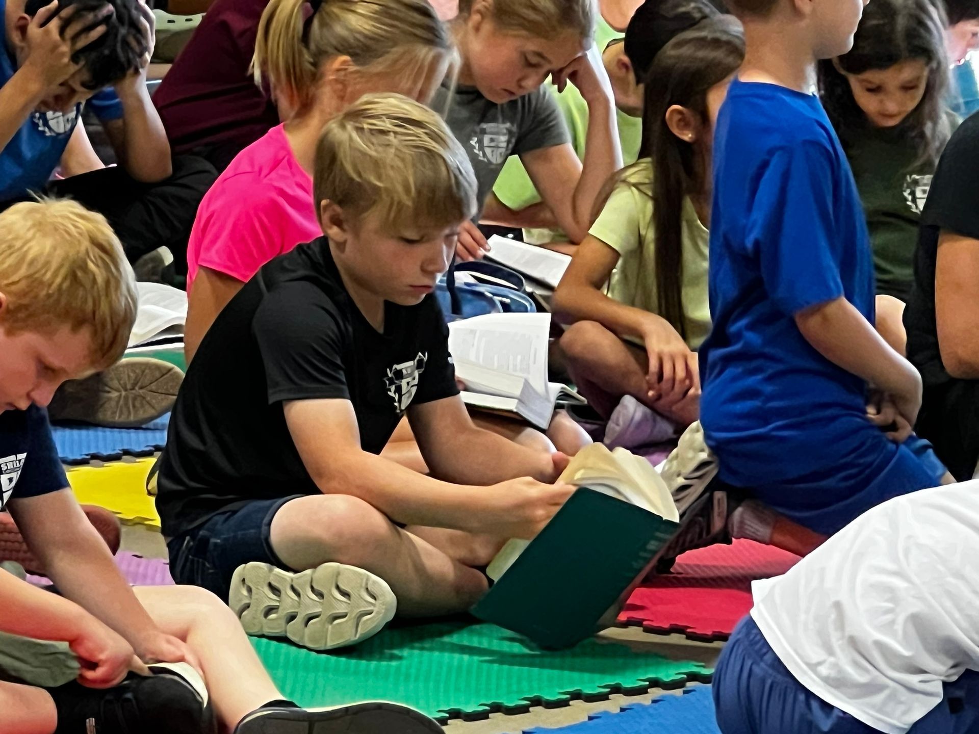 Children sitting on mats, reading books. A boy in black shirt focused on his book; other kids are also absorbed.