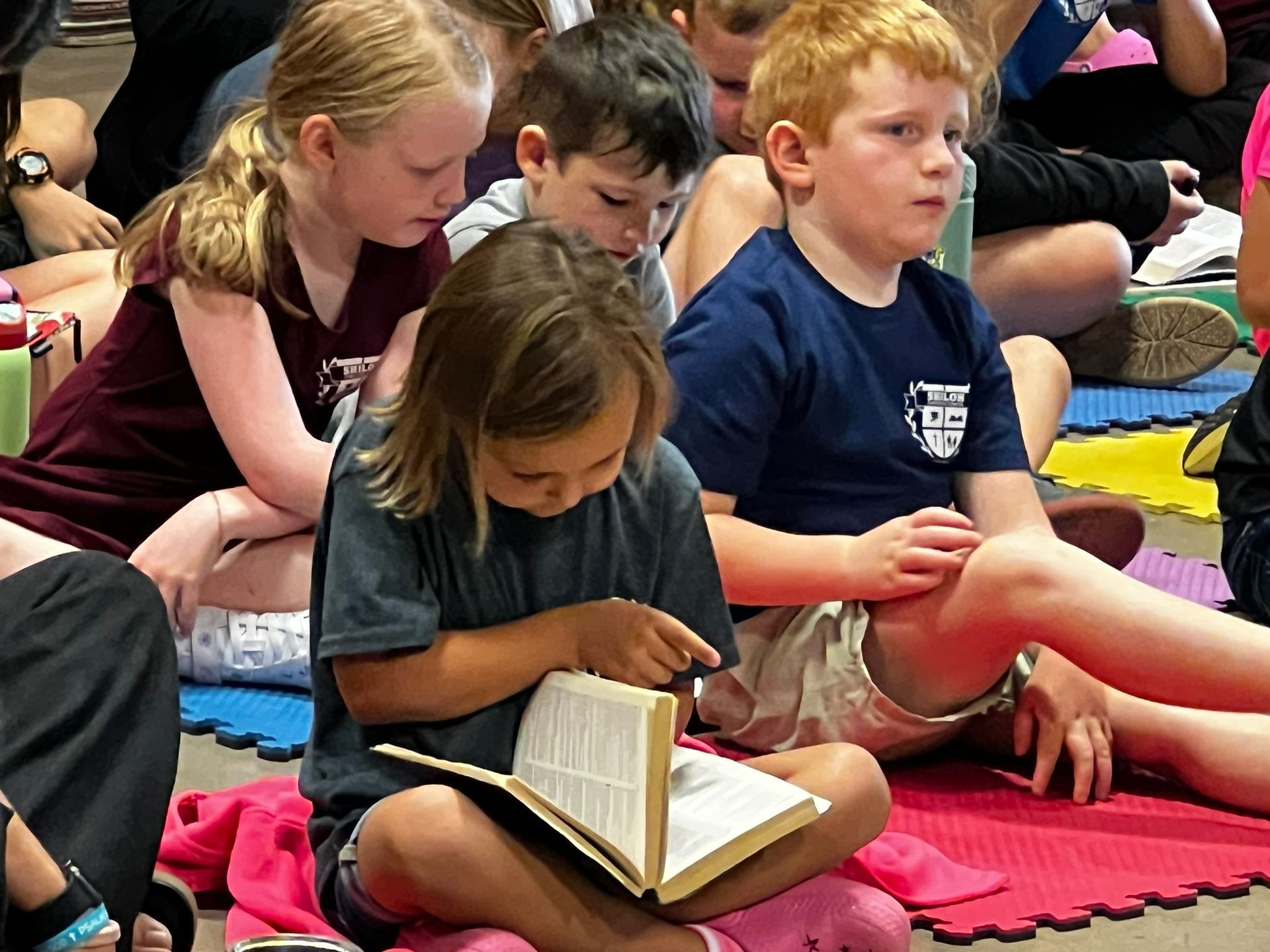 Children sitting on a floor, reading books. One girl points at her book.