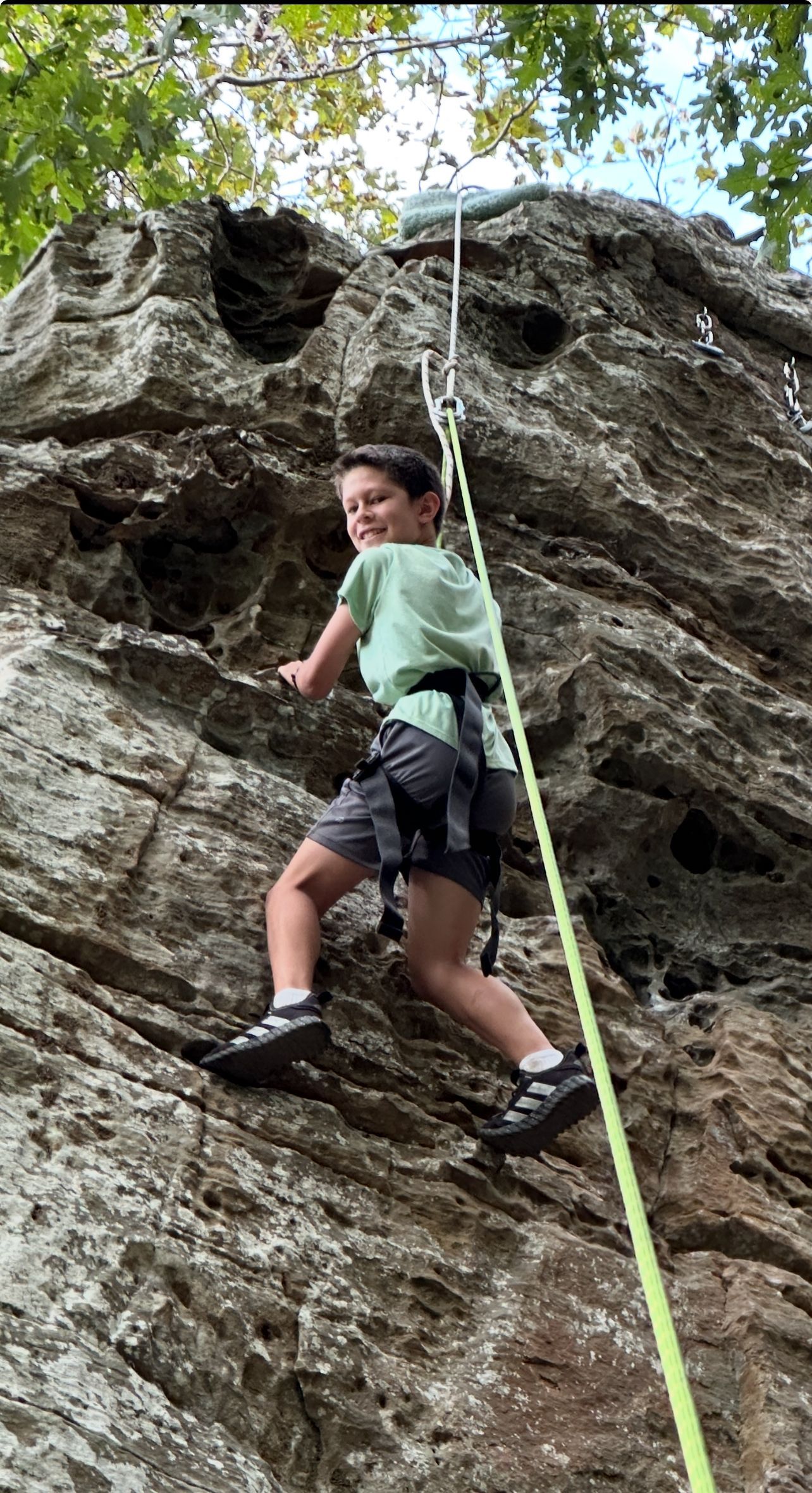 Boy smiles while rock climbing outdoors, wearing a harness and shoes, with a rope.
