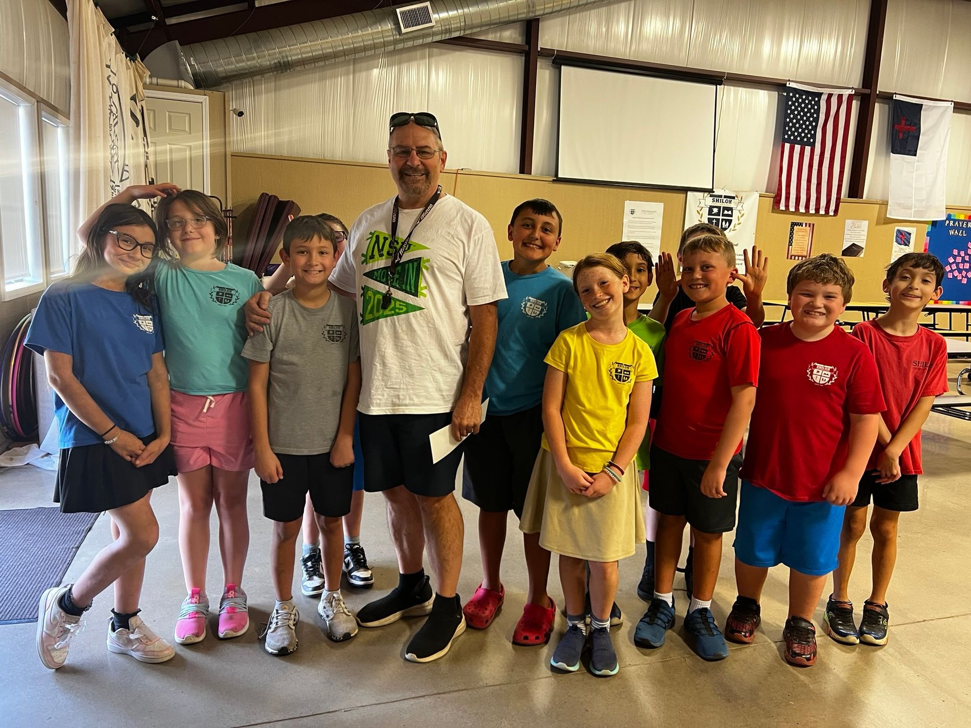 A group of children and a man stand indoors, smiling at the camera. They are wearing t-shirts, shorts, and sunglasses.