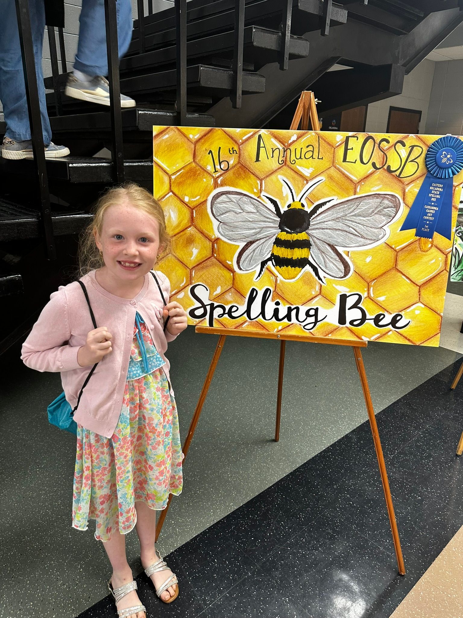 Smiling child beside a bee-themed sign on an easel, with a colorful outfit indoors
