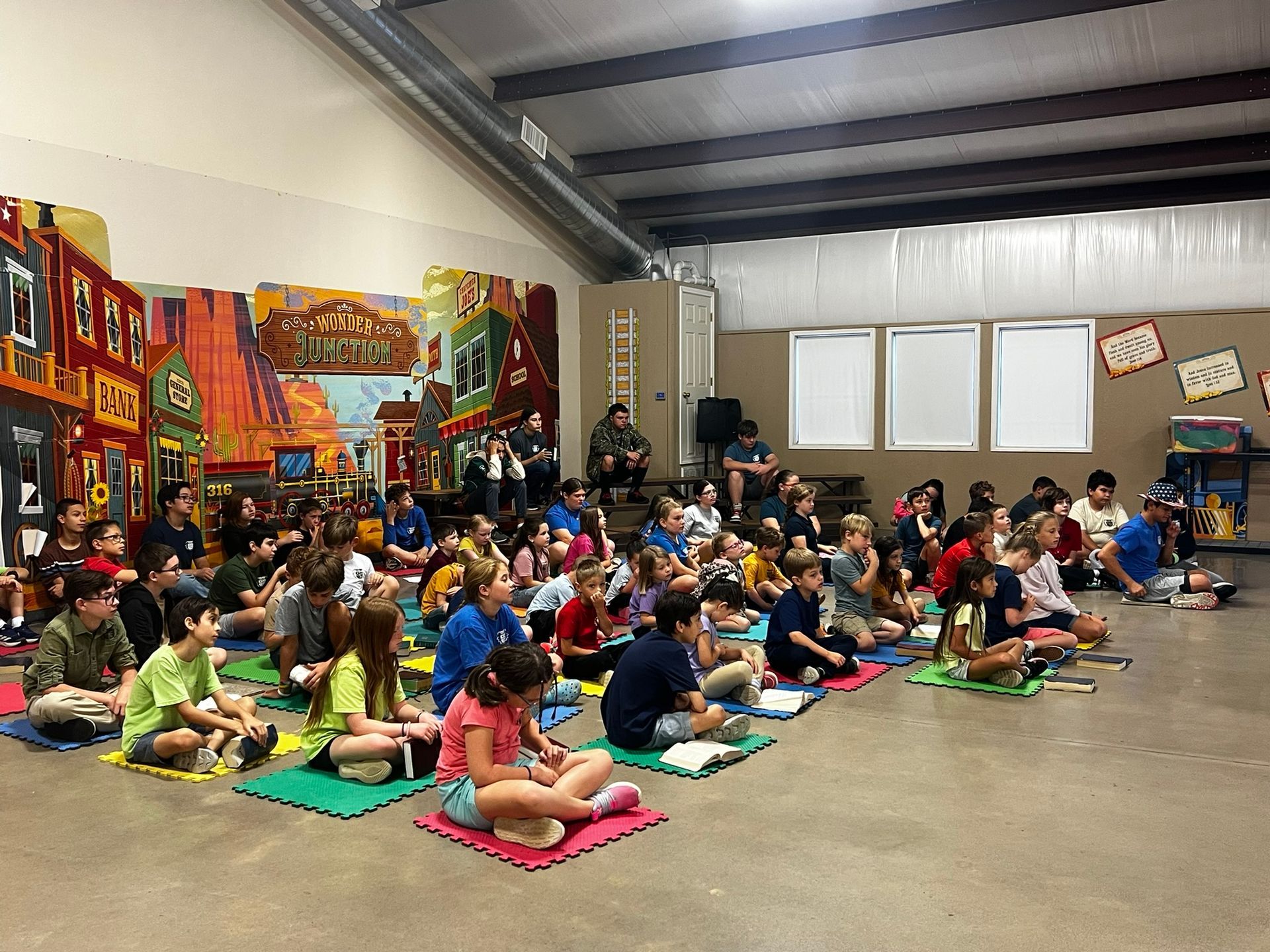 Children sitting on mats indoors, watching presentation. Colorful mural on wall.