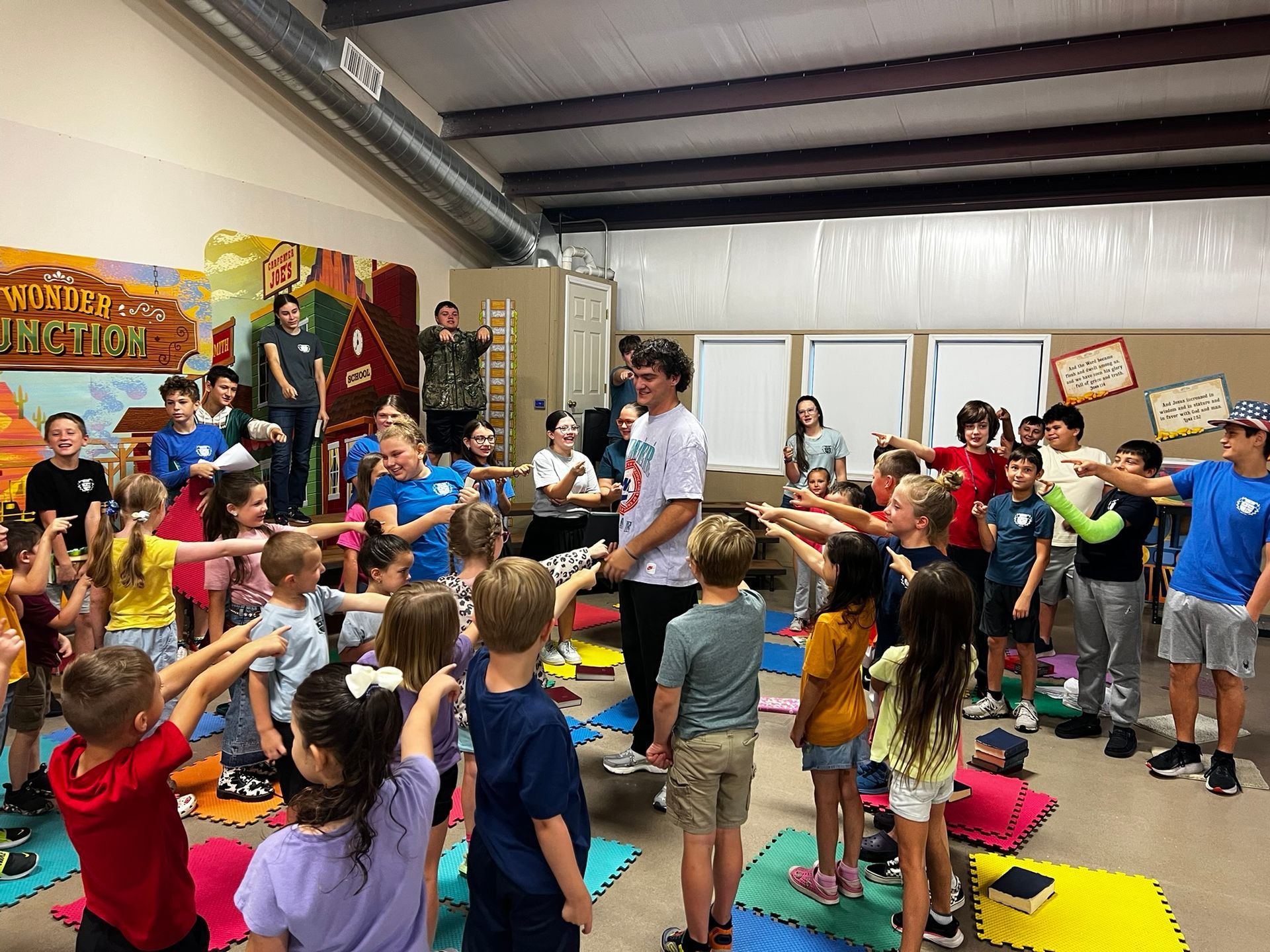 Kids point at a man in a room with painted murals. Others watch.