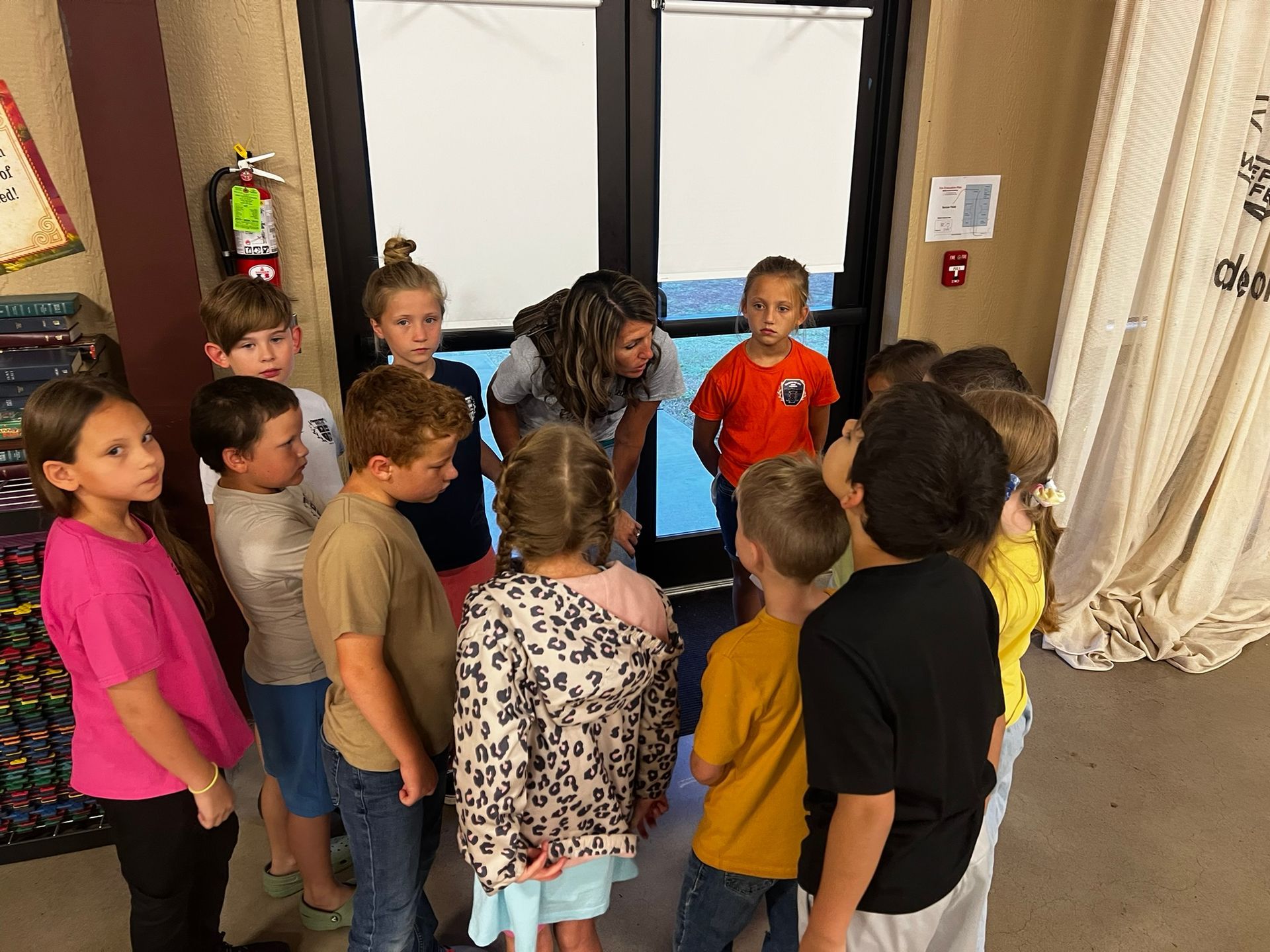 Children and teacher in a circle, looking down at something, near glass doors.