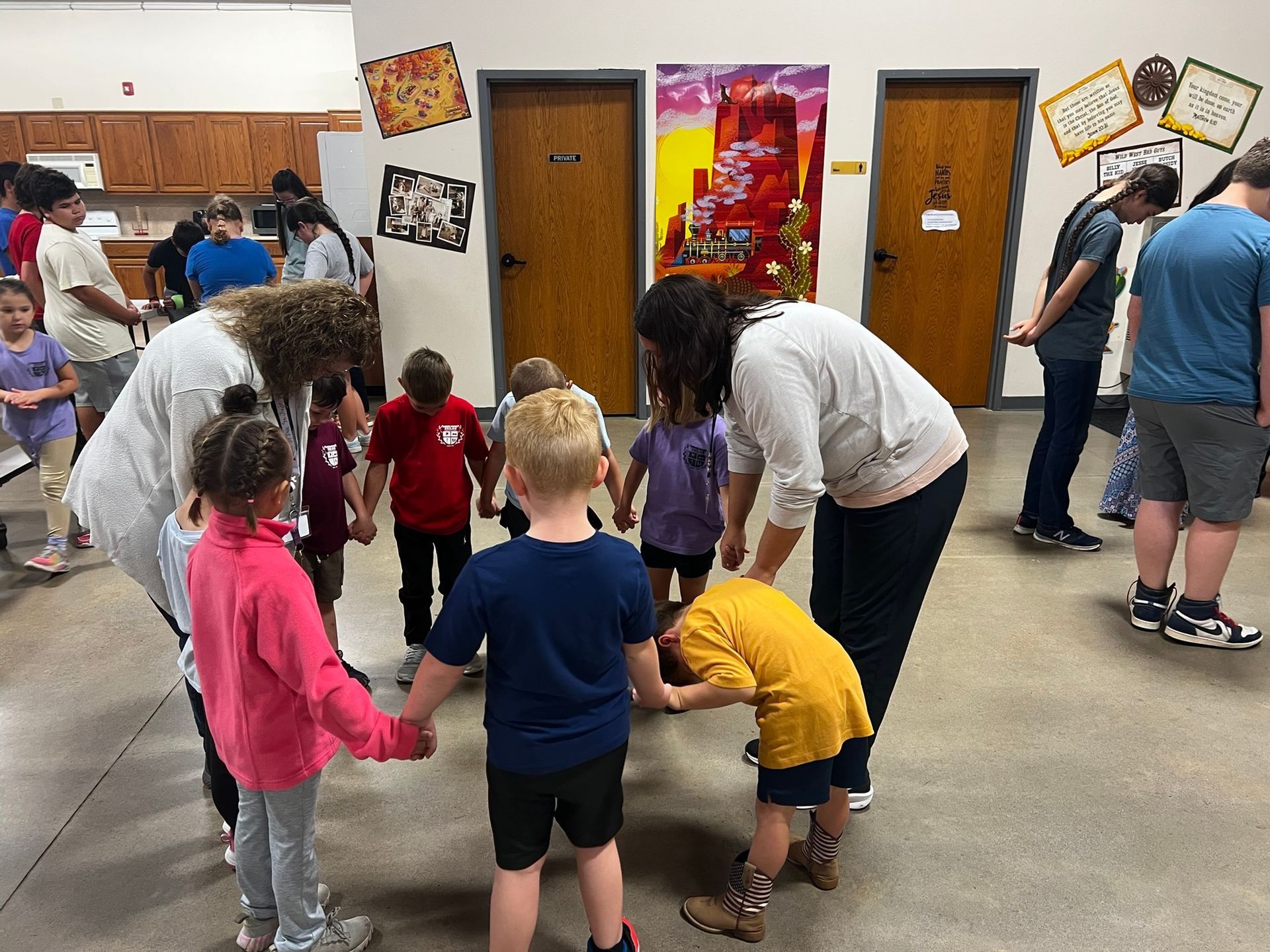 Children and adults in a circle, holding hands. A teacher guides them. Indoors, wood doors and colorful art on the walls.