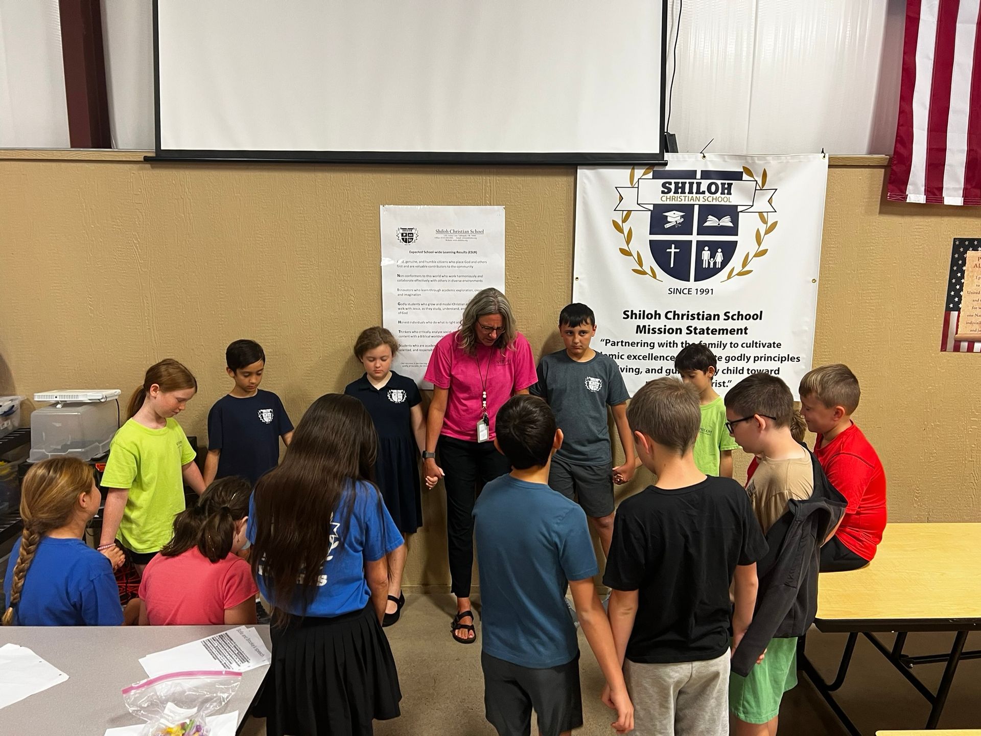 Group of students and teacher holding hands in a circle, indoors near a banner and a screen.