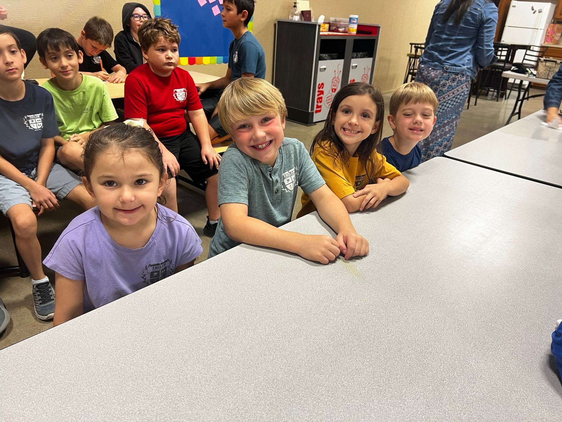 Children smiling at a table in a classroom. Others sit nearby.