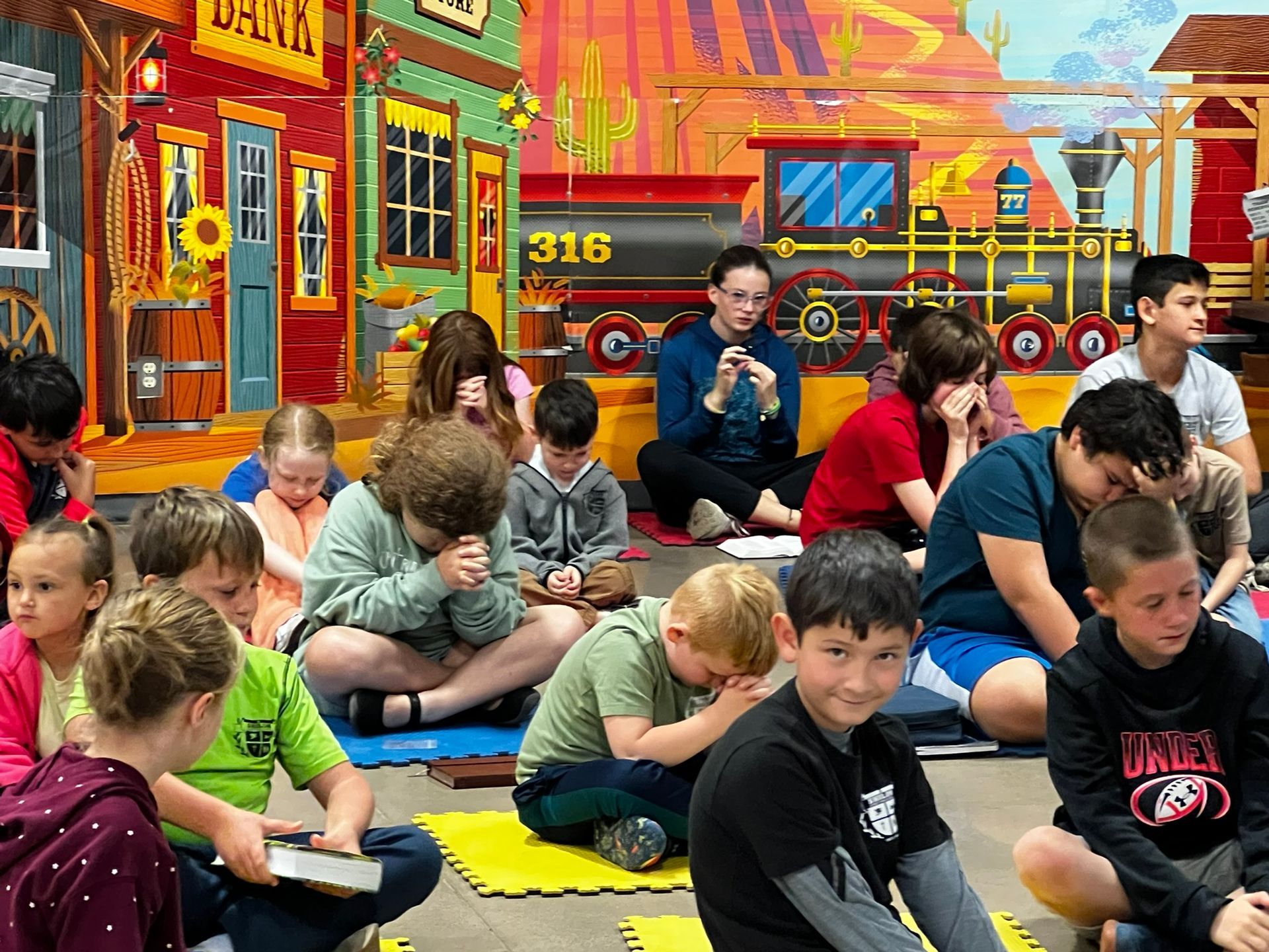 Children and an adult kneeling in prayer, in a classroom with a western-themed mural.