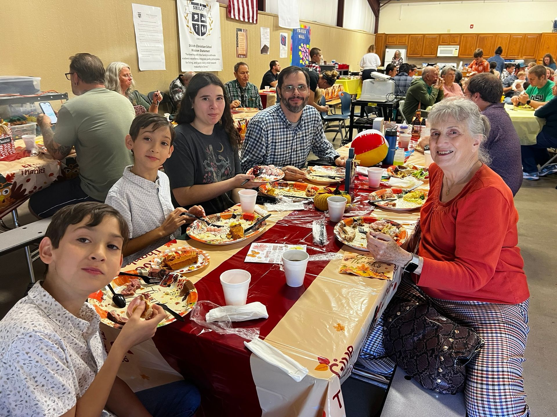 Family smiles while eating at a long table in a community hall, autumn-themed decor.