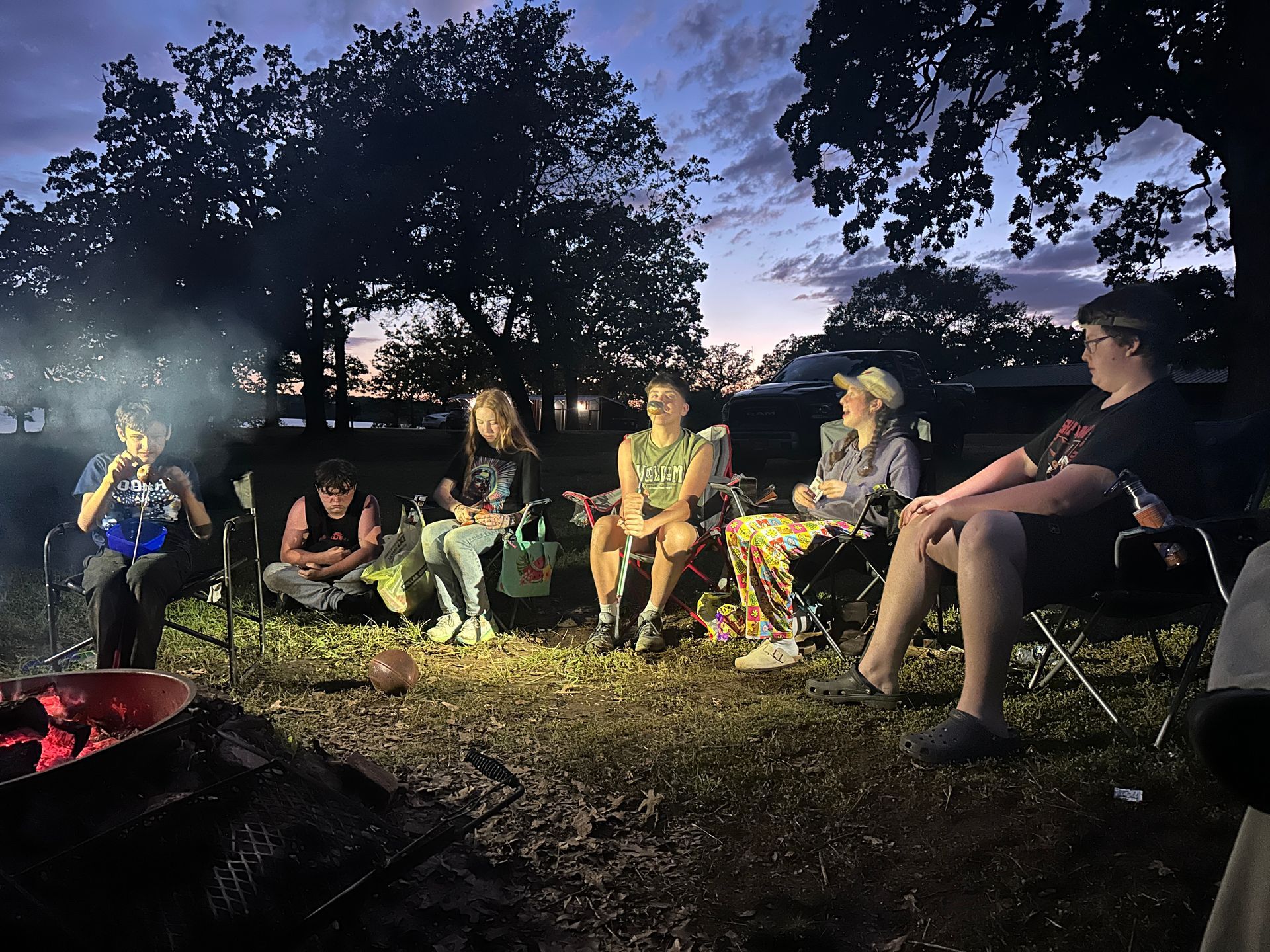 Group of people around a campfire at dusk, talking and enjoying the evening outdoors.