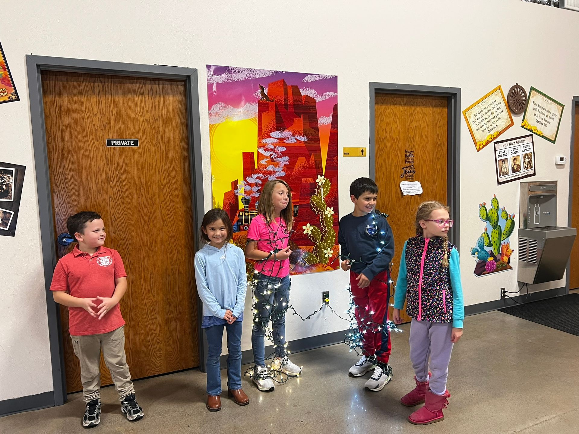 Five children stand in a school hallway with a colorful painting on the wall.