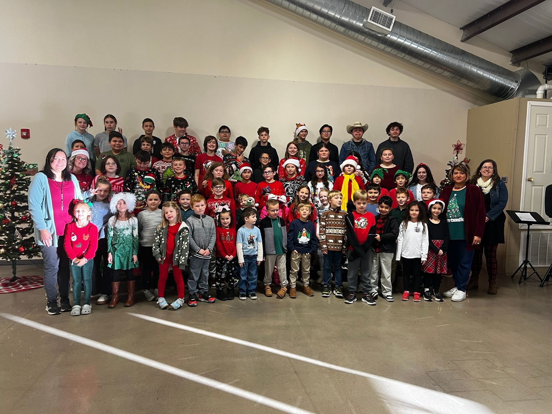 Group of people, mostly children, in a room with a Christmas tree. Many wearing holiday sweaters.
