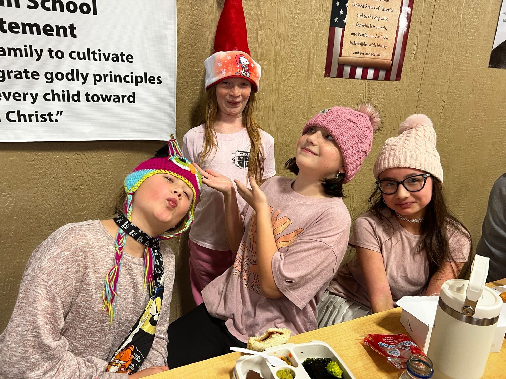 Four girls wearing hats, posing playfully at a table, likely in a school or event setting.