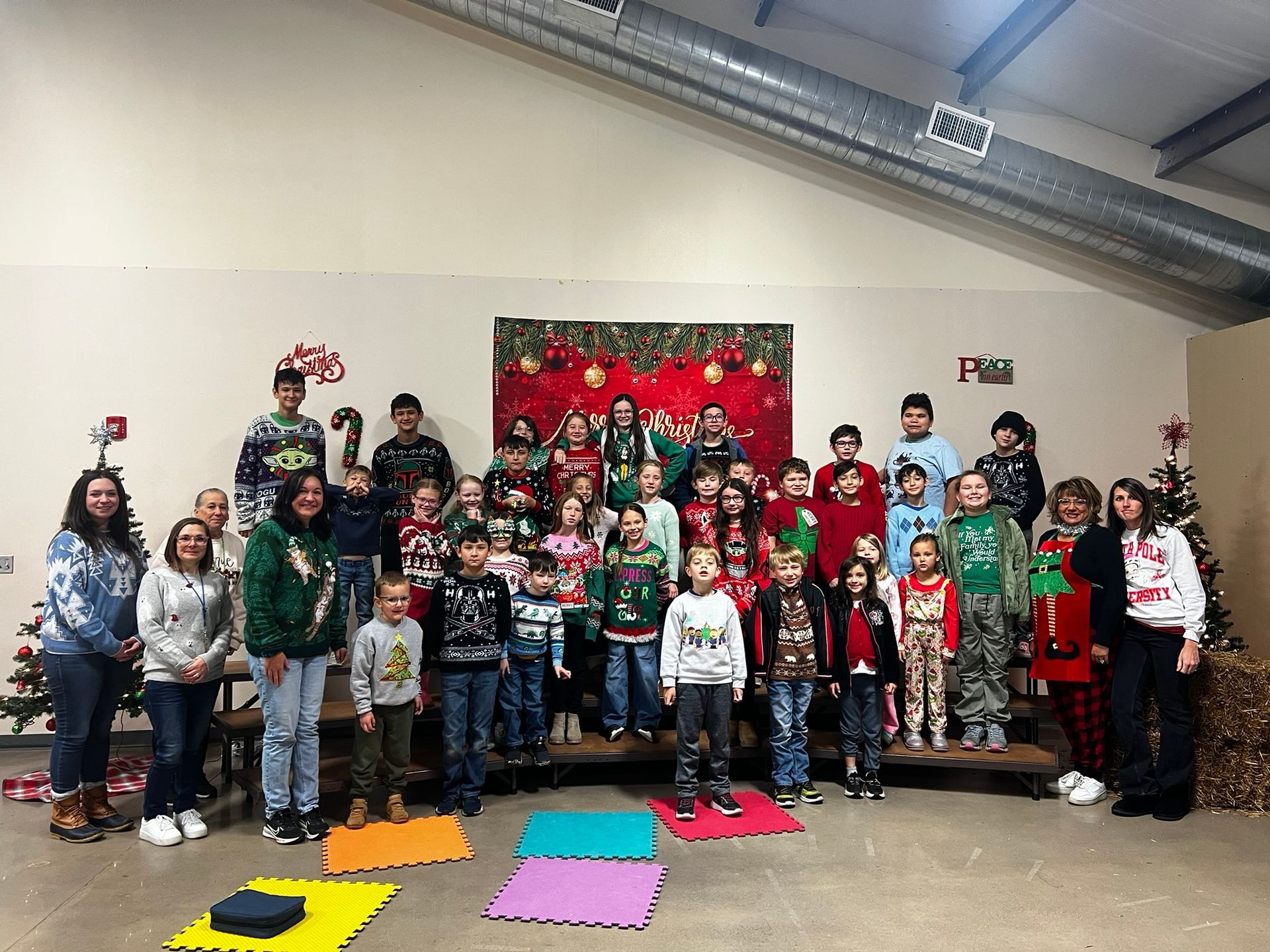 Group of children in holiday sweaters standing on stage with adults and decorations.
