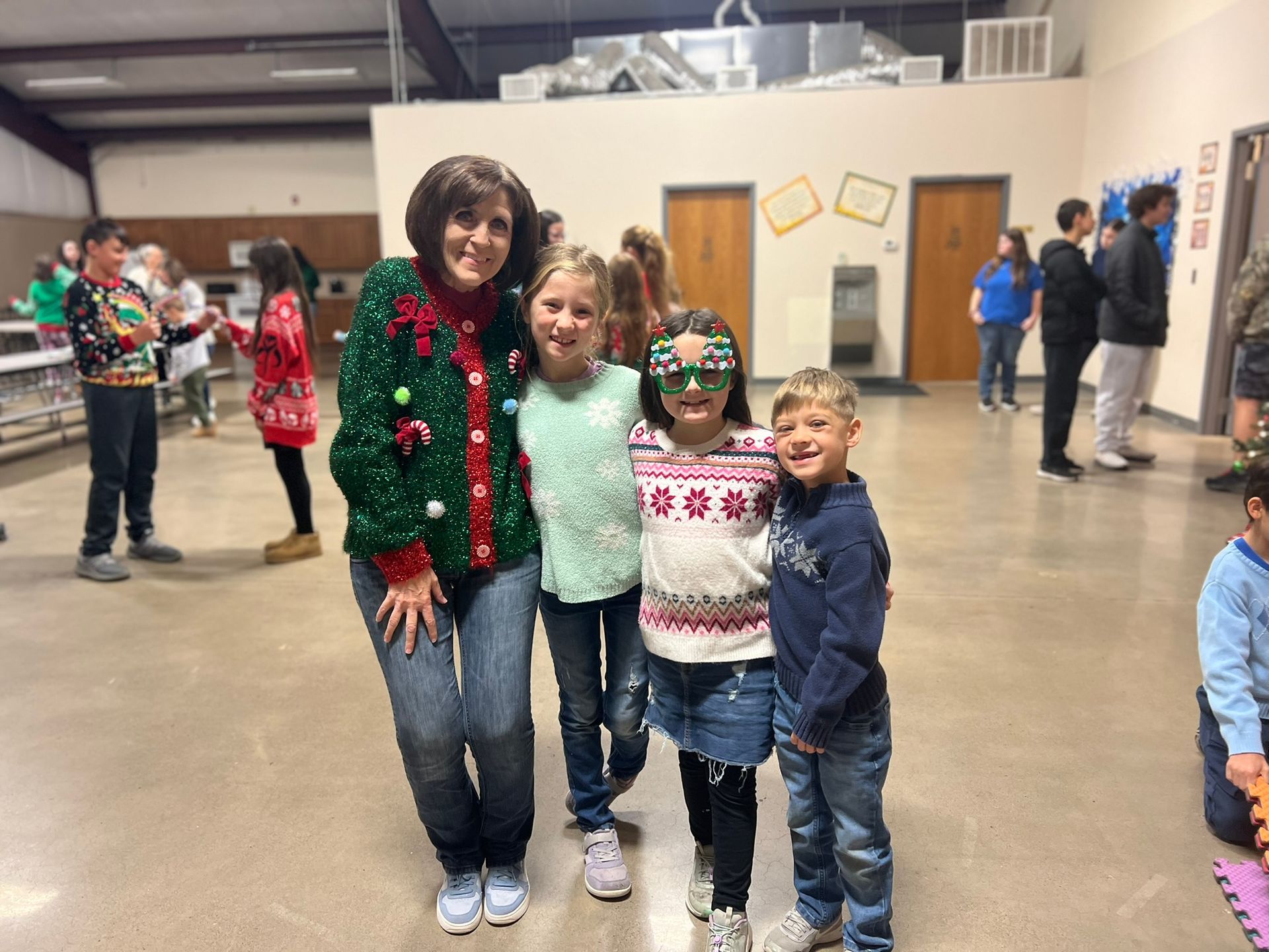 Woman in festive sweater poses with three children in a school setting; people in background.