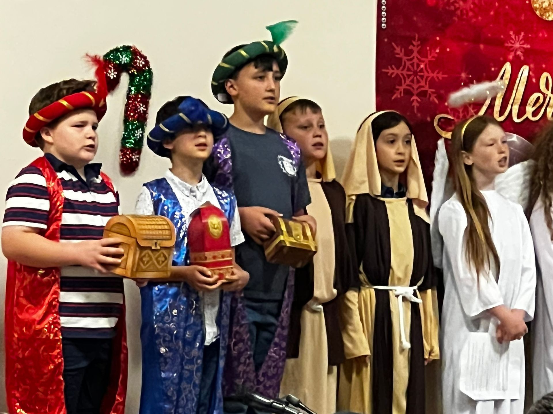 Children in Christmas play costumes holding gifts, singing near a Christmas backdrop.