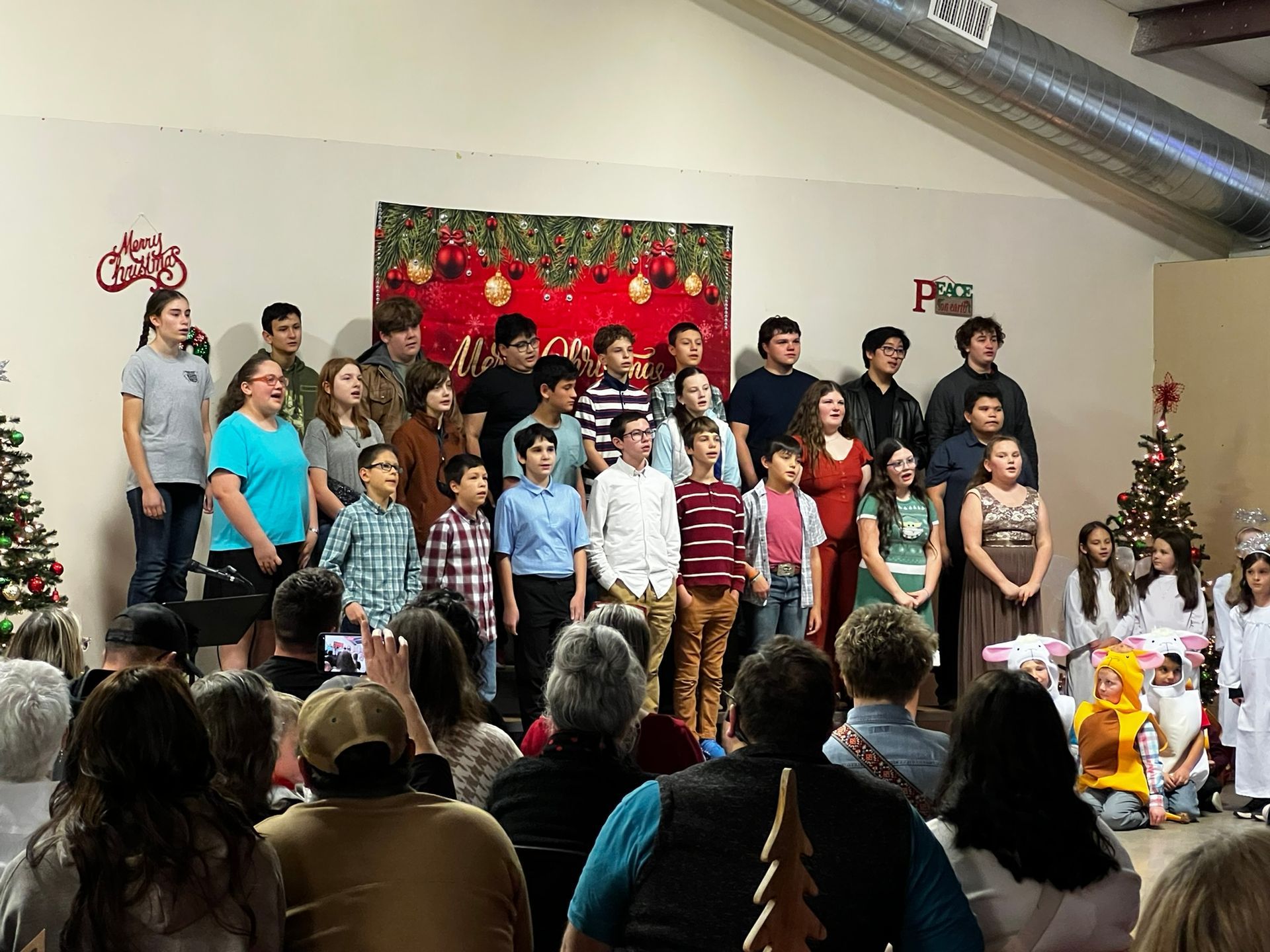 Children's holiday performance onstage, audience watching. Red and green decorations, Christmas trees.