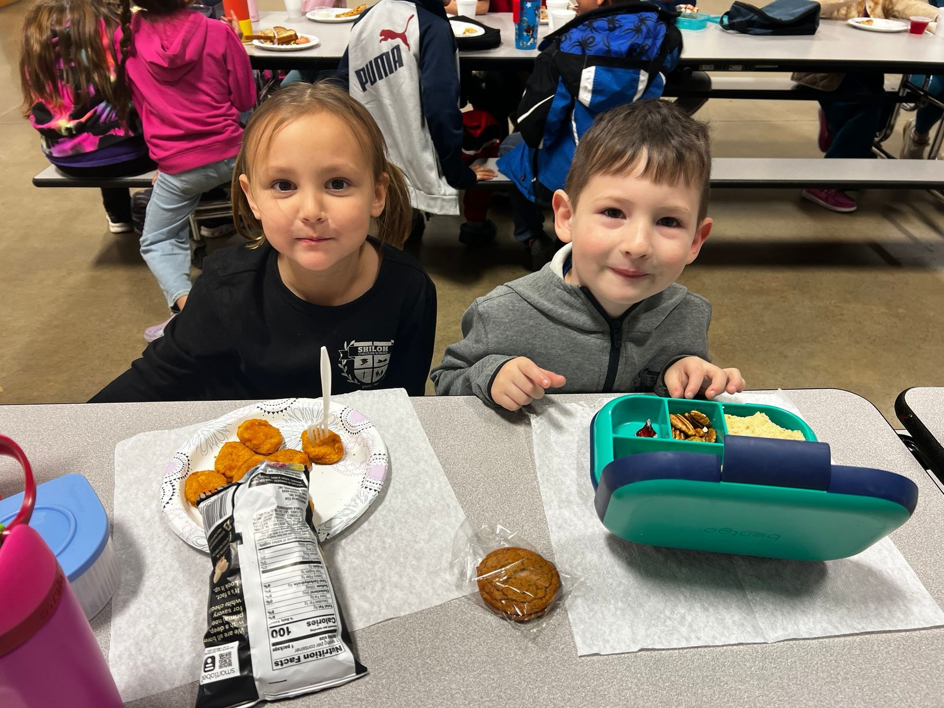 Two children smiling at a table with lunch: chicken nuggets, a cookie, and a boat-shaped lunchbox.
