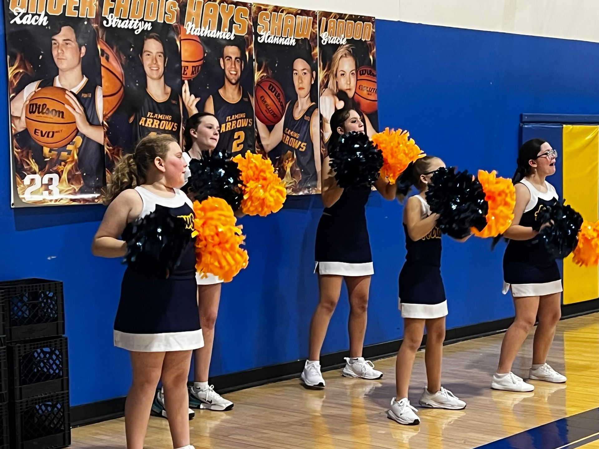 Cheerleaders in navy and white uniforms with orange and black pom-poms perform in a gym, with basketball player banners in the background.