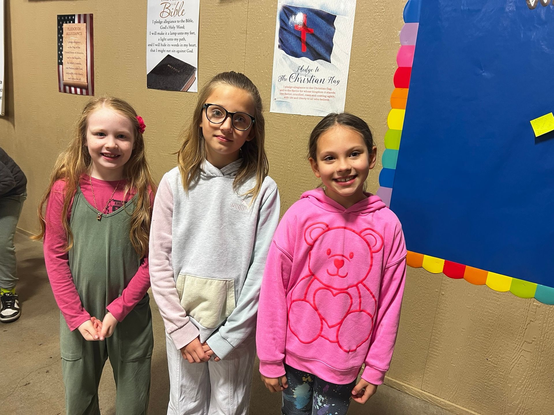 Three young girls smiling in front of a bulletin board, wearing colorful casual clothes.