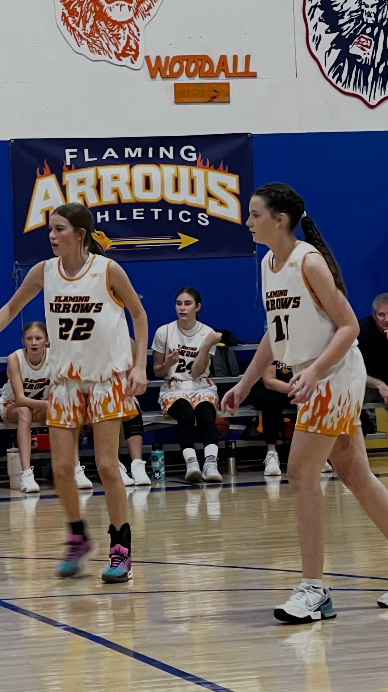 Two basketball players in white and orange uniforms on court, Flaming Arrows banner in background.