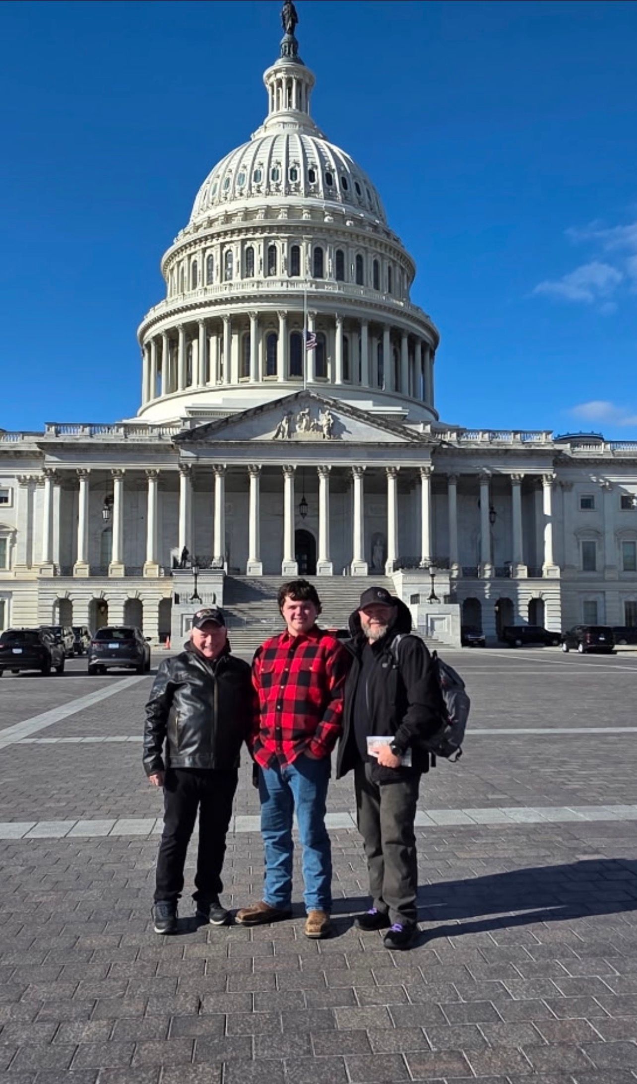 Three people pose in front of the US Capitol Building on a clear day.