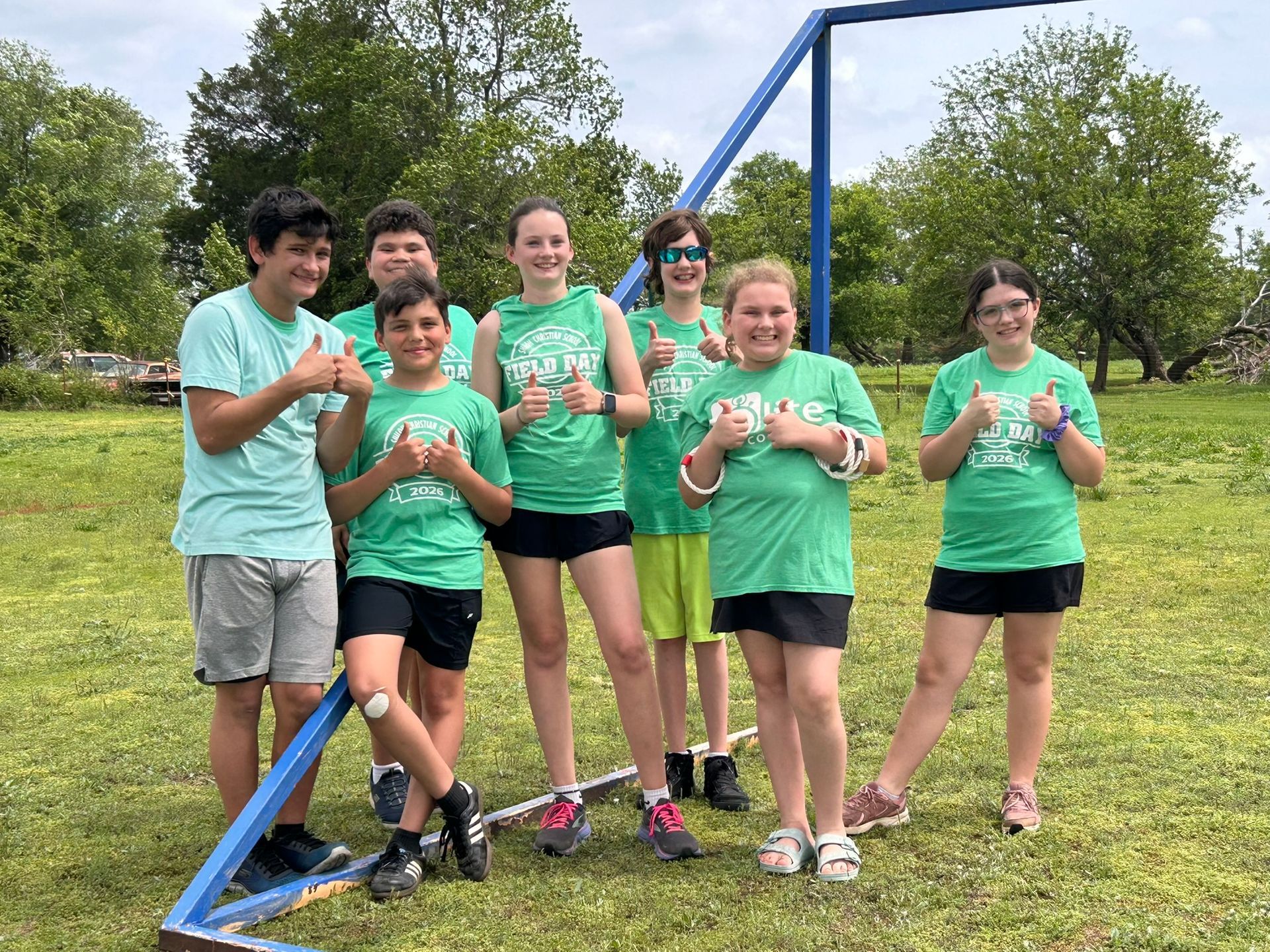 Six people in green shirts pose outdoors beside a blue metal frame on grass.